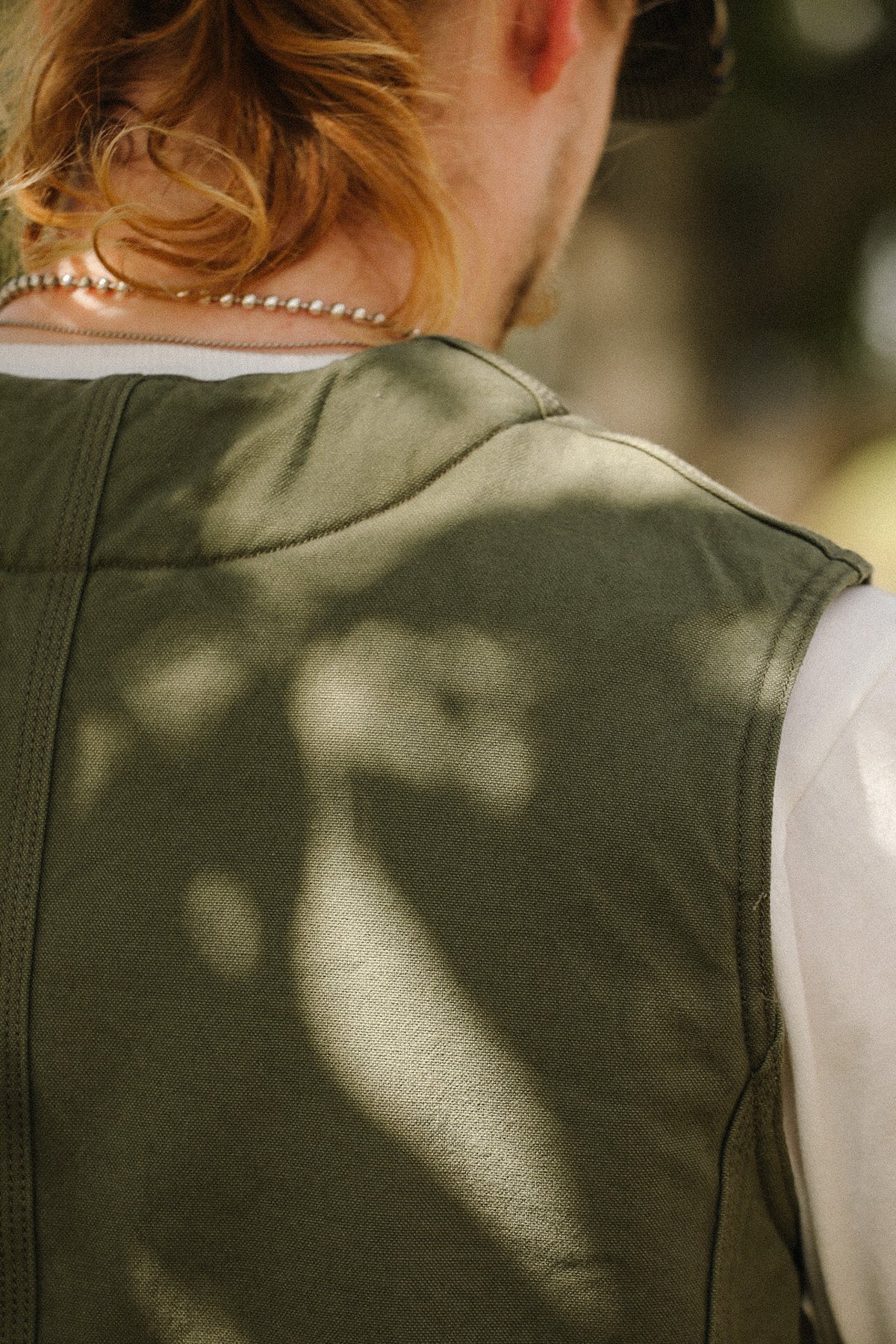 Close-up back view of a person wearing an olive green sleeveless vest over a white shirt, with dappled sunlight and leaf shadows across the fabric.