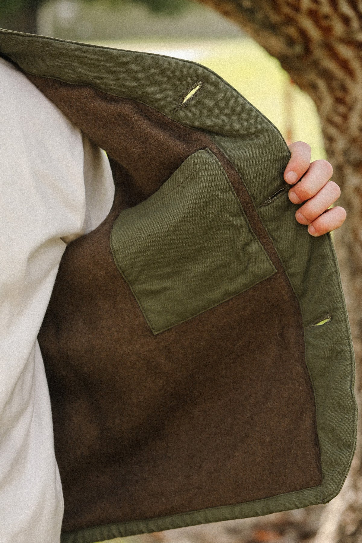Close-up of a person holding open an olive green jacket to show a brown wool-like lining and a large patch pocket, with a blurred outdoor background.