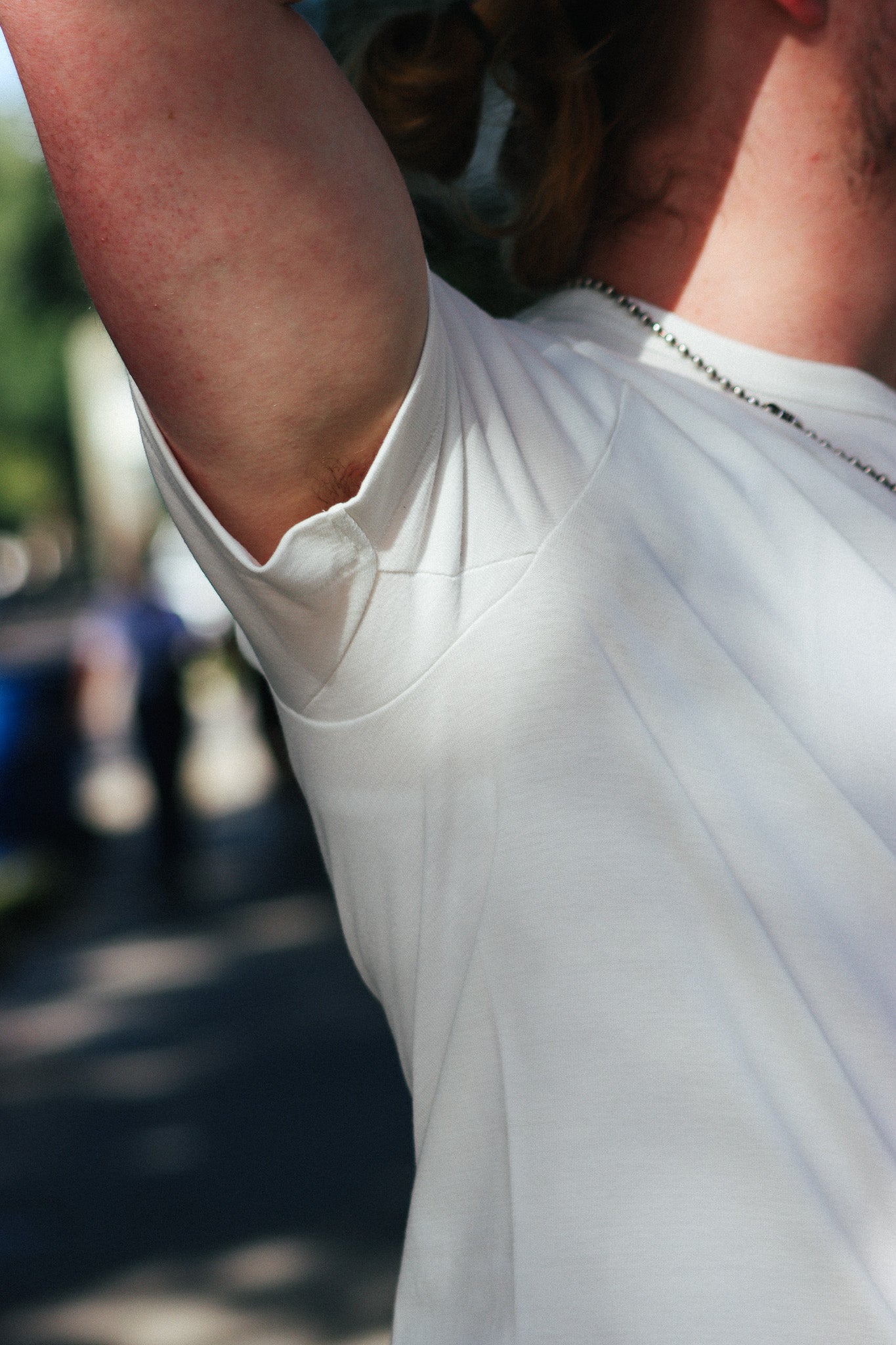 Close-up of a person wearing a white short-sleeve t-shirt outdoors, showing the shoulder, arm, neck, and upper chest with a blurred street background.