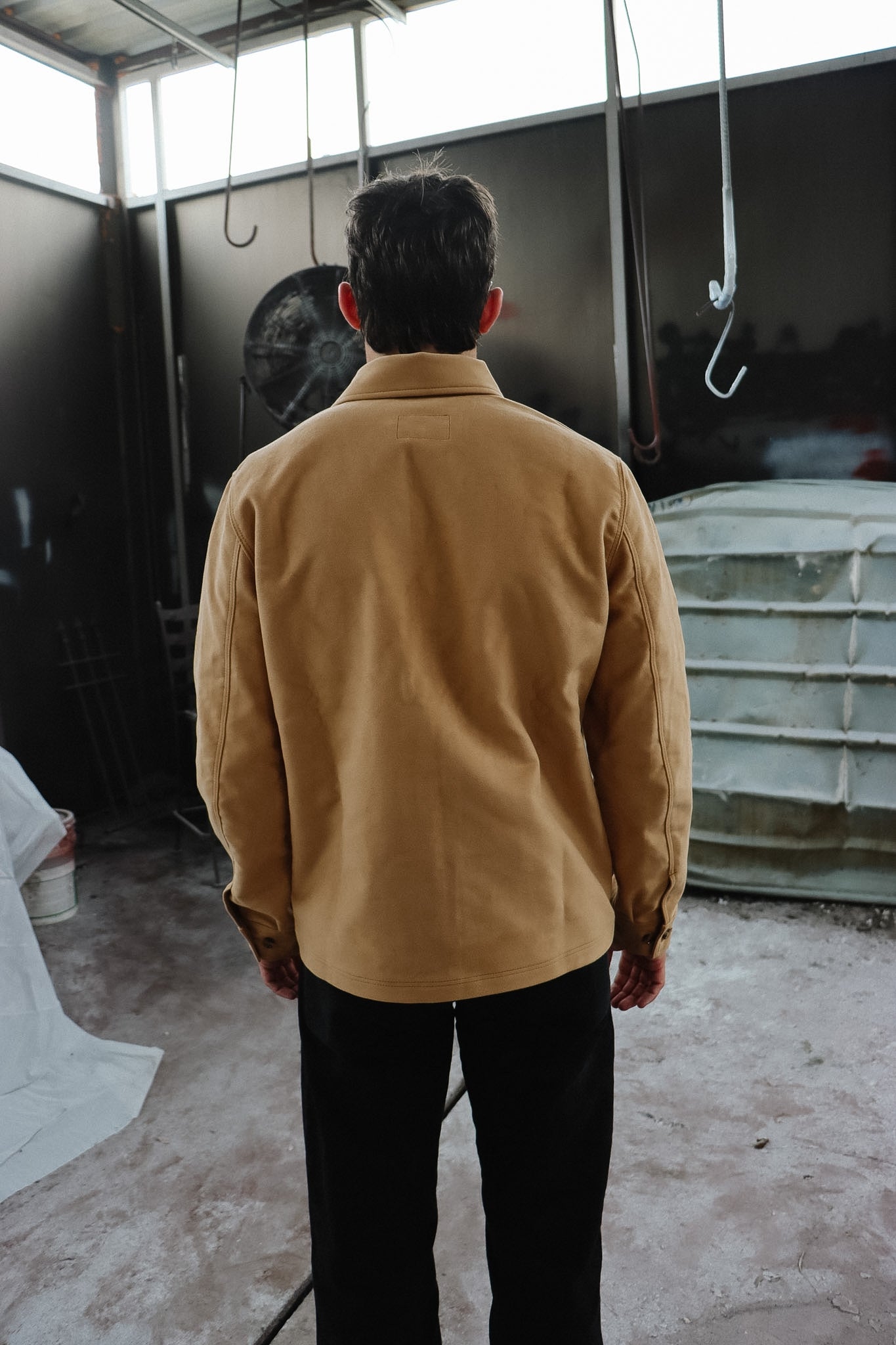 Person wearing a tan jacket standing in an industrial setting with hooks and a dark wall.
