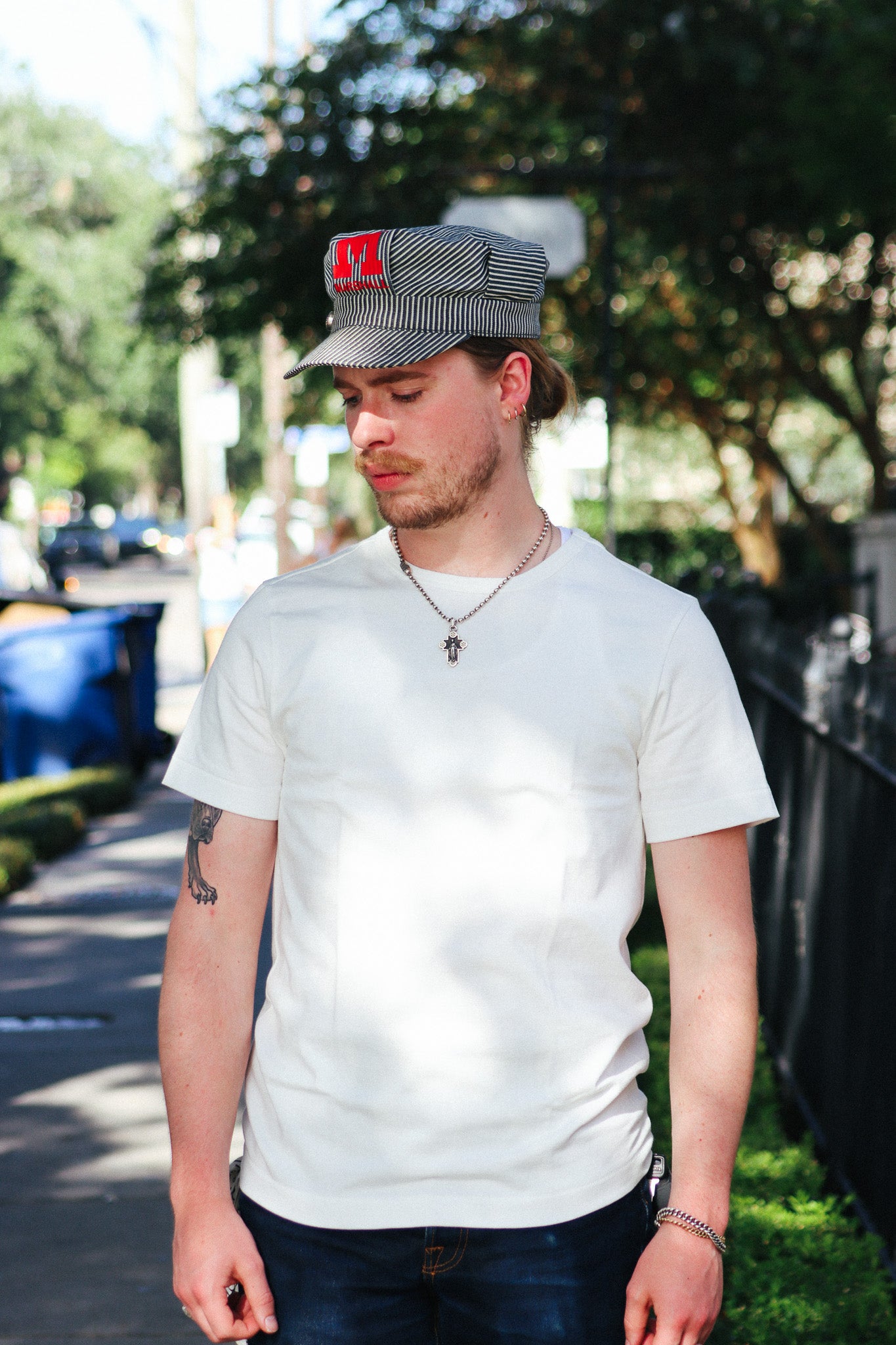 A man wearing a white crewneck t-shirt, dark blue jeans, a gray and black striped cap with a red 'H' logo, and a silver necklace, standing outdoors on a sunny sidewalk.