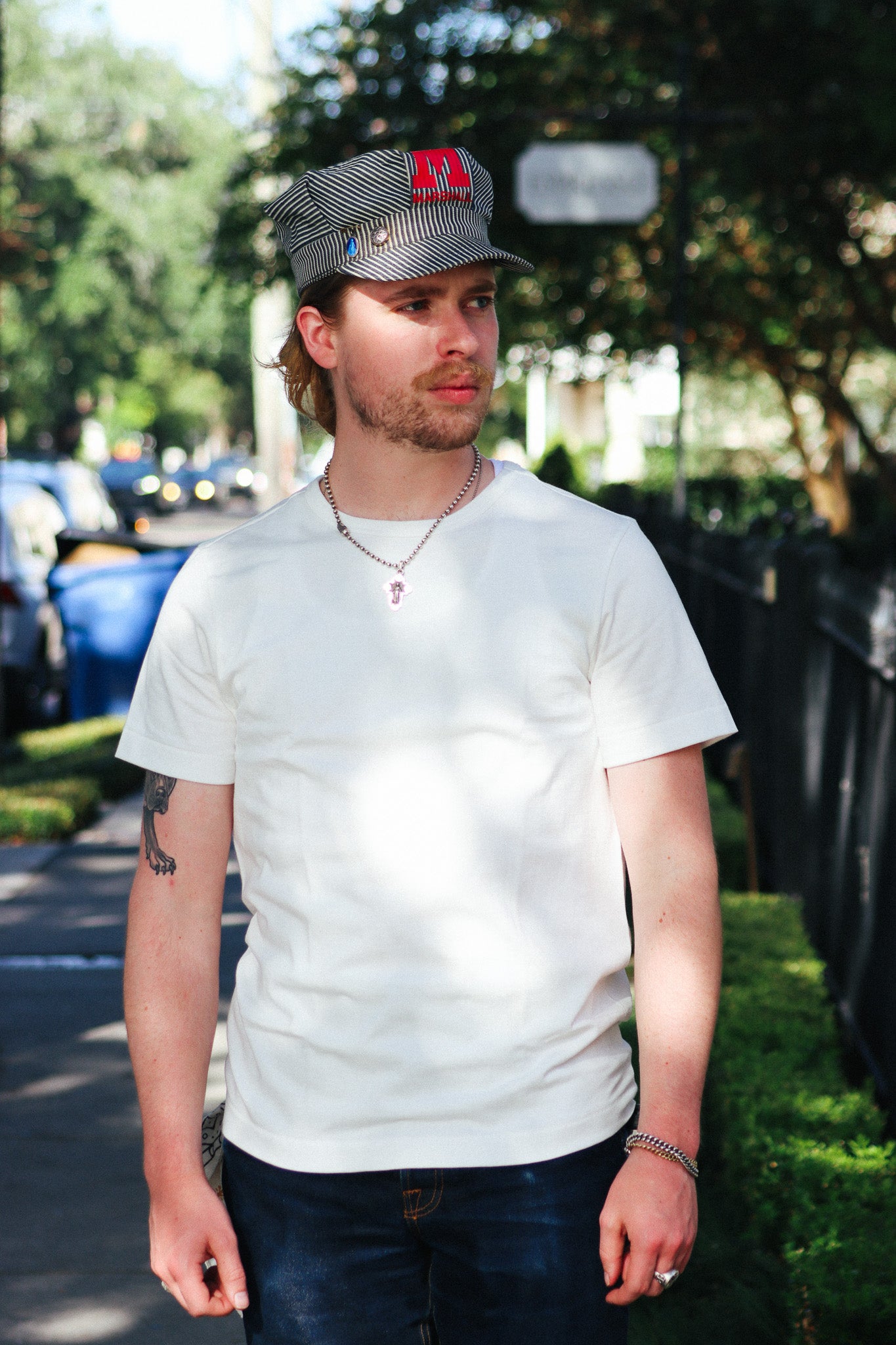 A man wearing a white crewneck t-shirt, dark blue jeans, a gray and white checkered cap with a red logo, and a silver chain necklace, standing outdoors on a sunny day.