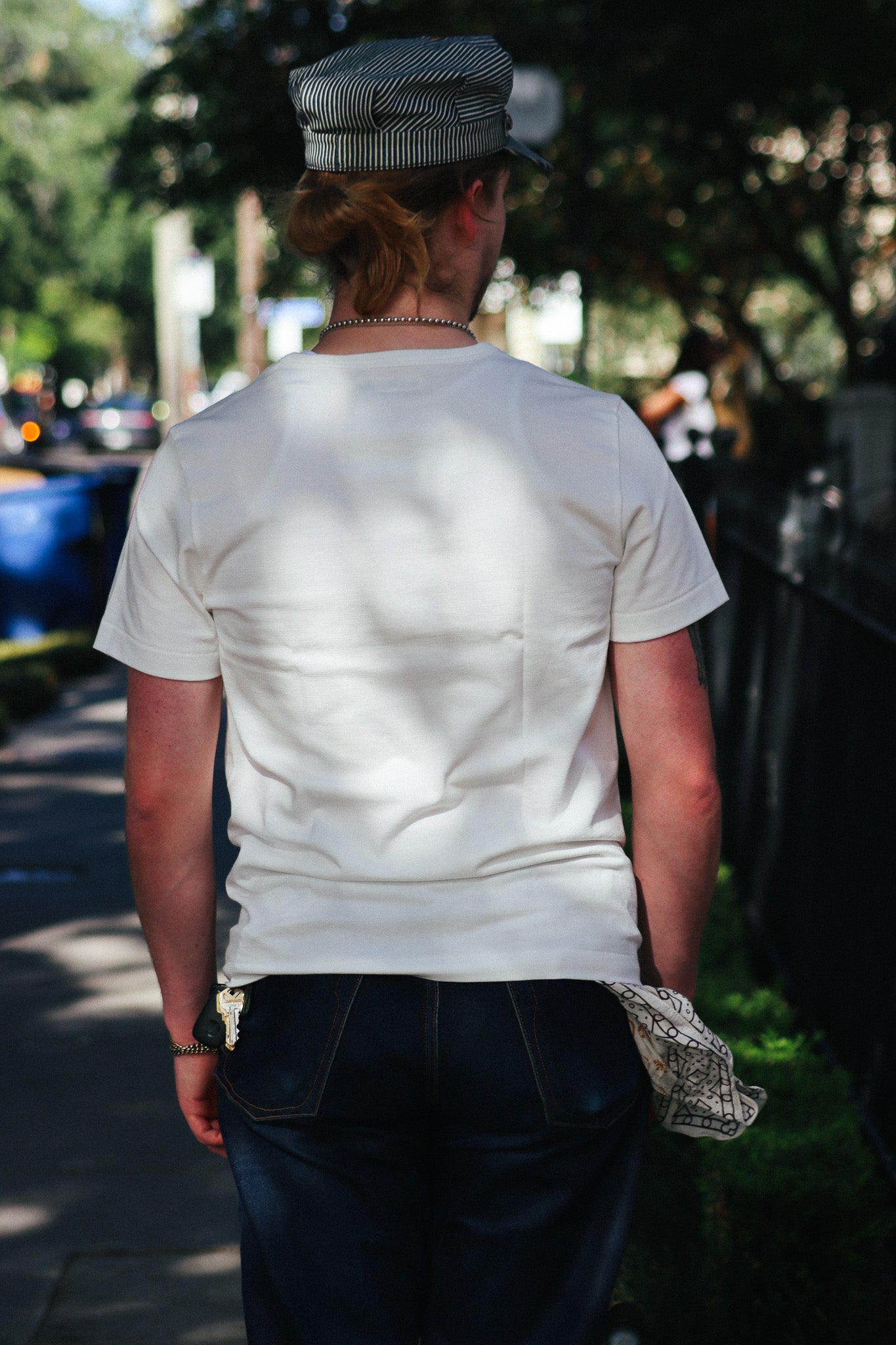 A person viewed from behind wearing a white short-sleeve crewneck t-shirt, dark blue jeans, and a gray and white striped cap, walking outdoors on a sunny sidewalk.