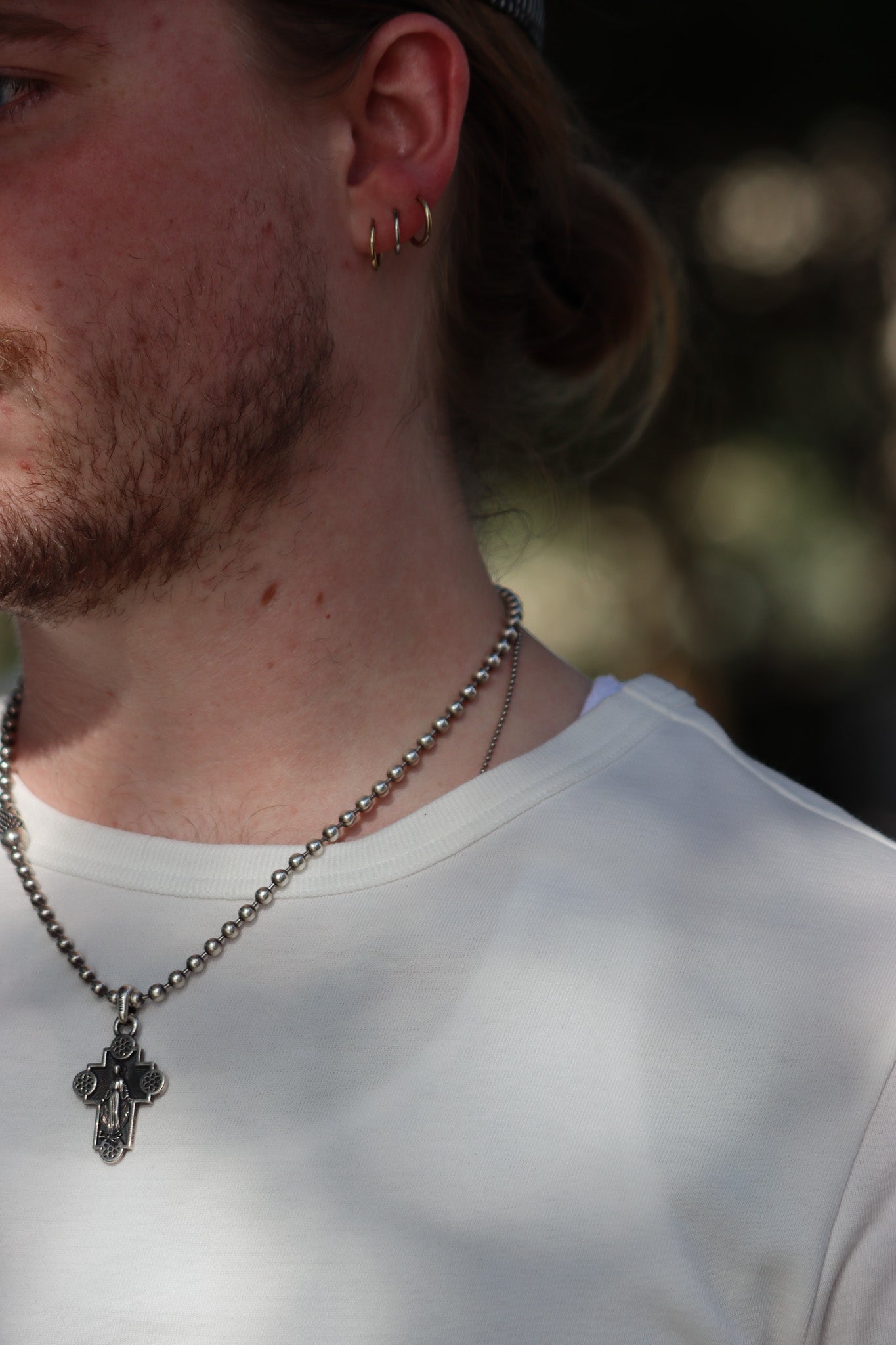 A close-up lifestyle shot of a man wearing a silver ball chain necklace with a cross pendant, along with gold hoop earrings and a white t-shirt.