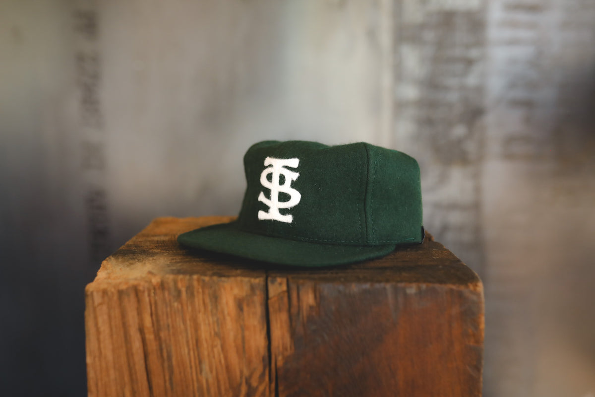 A dark green snapback cap with a white embroidered monogram logo on the front, displayed on a rustic wooden block against a blurred gray background.