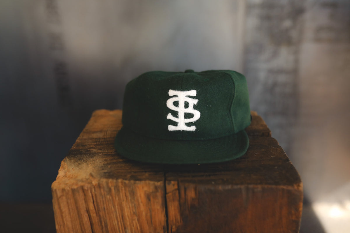 A dark green flat-brim cap with a white embroidered monogram logo on the front, displayed on a rustic wooden block against a blurred gray background.