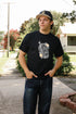Young man wearing black Iron Shop Provisions coffin graphic tee and dark jeans outdoors on sidewalk
