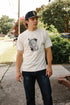 Young man wearing cream graphic t-shirt with coffin design and black baseball cap, standing on sidewalk with greenery background