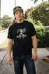 Young man wearing black loopwheel tee with vintage race car and slogan graphic, outdoors on sidewalk