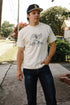 Man wearing cream Iron Shop Provisions Buffalo Nickel Starting Line Loopwheel tee and black cap standing on sidewalk