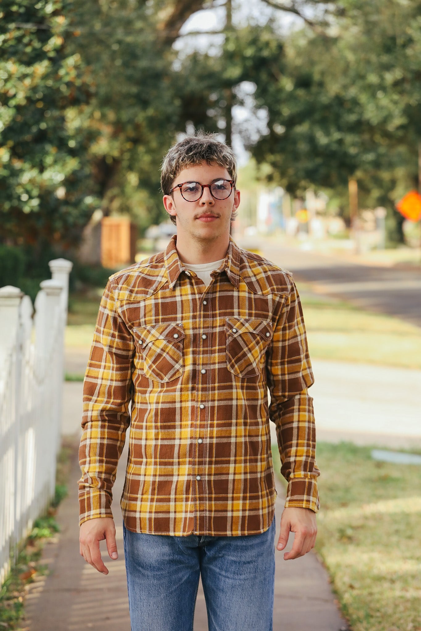 Person wearing a plaid shirt and glasses standing outdoors on a sidewalk.