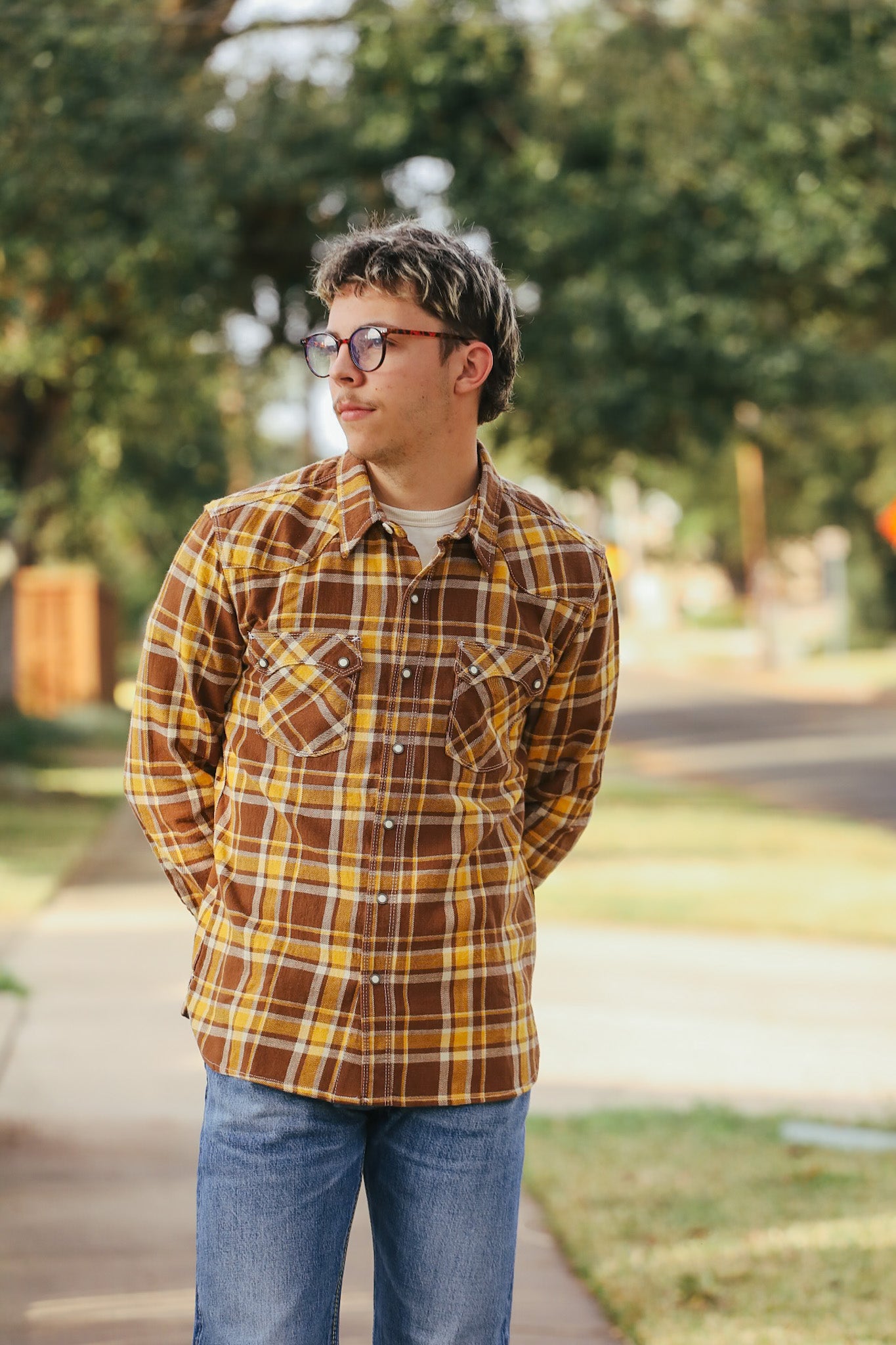 Person wearing a plaid shirt and glasses standing outdoors with trees in the background