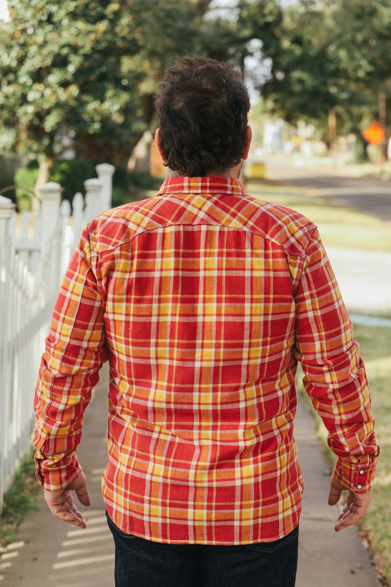 Person wearing a red and yellow plaid shirt walking on a sidewalk with trees and a white fence in the background.