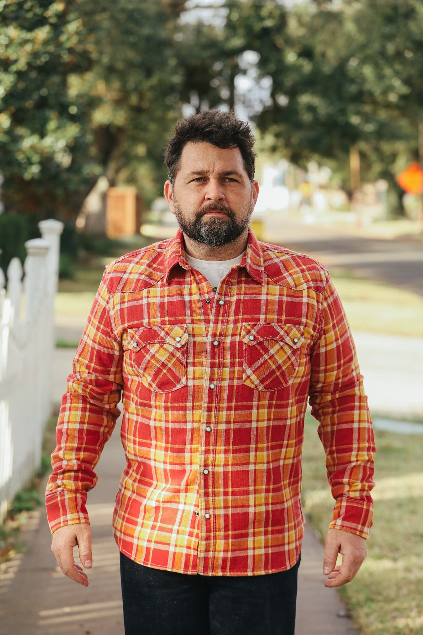 Man wearing a red and yellow plaid shirt standing outdoors with trees and a road in the background.