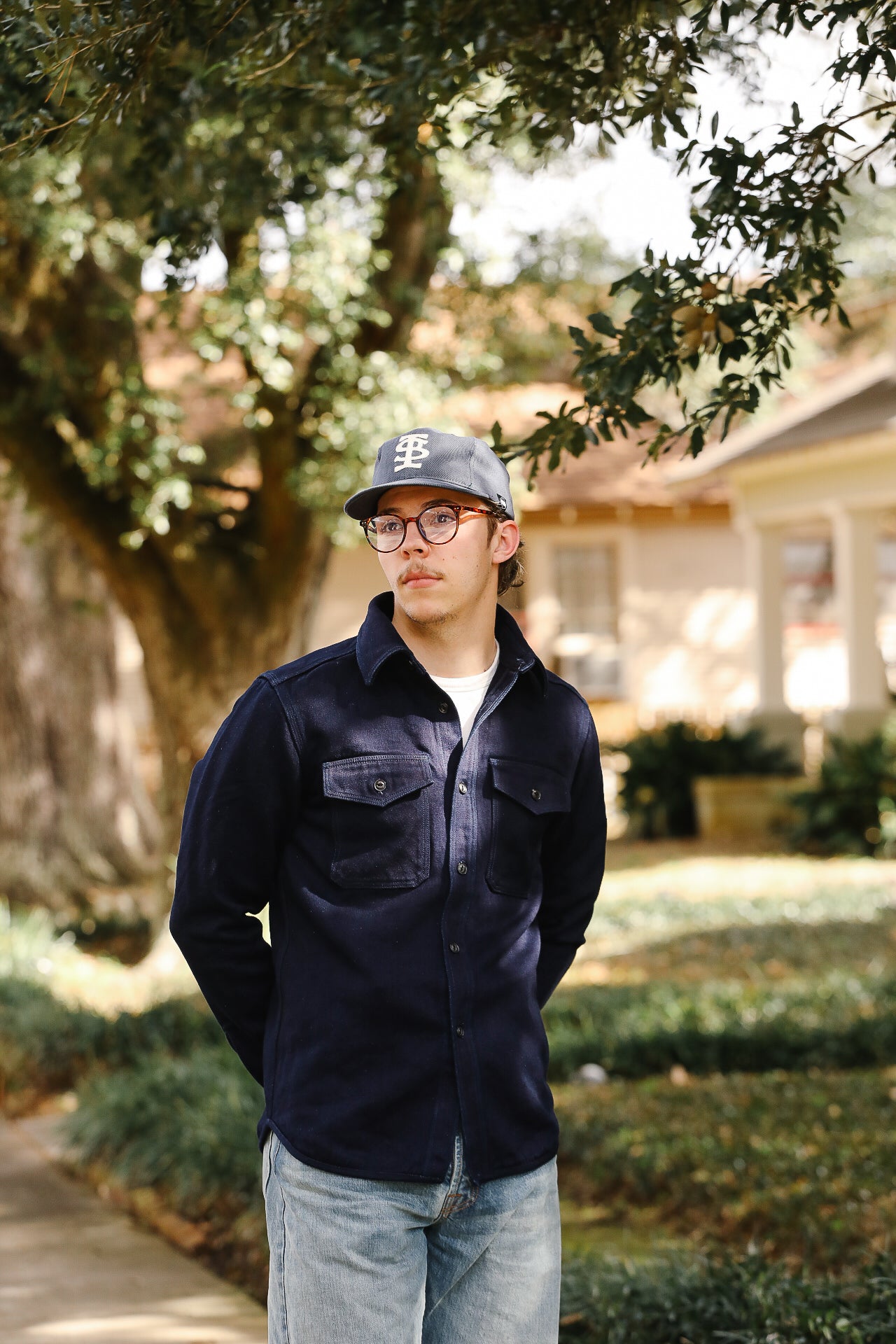 Person wearing a cap and glasses standing outdoors with trees and a house in the background