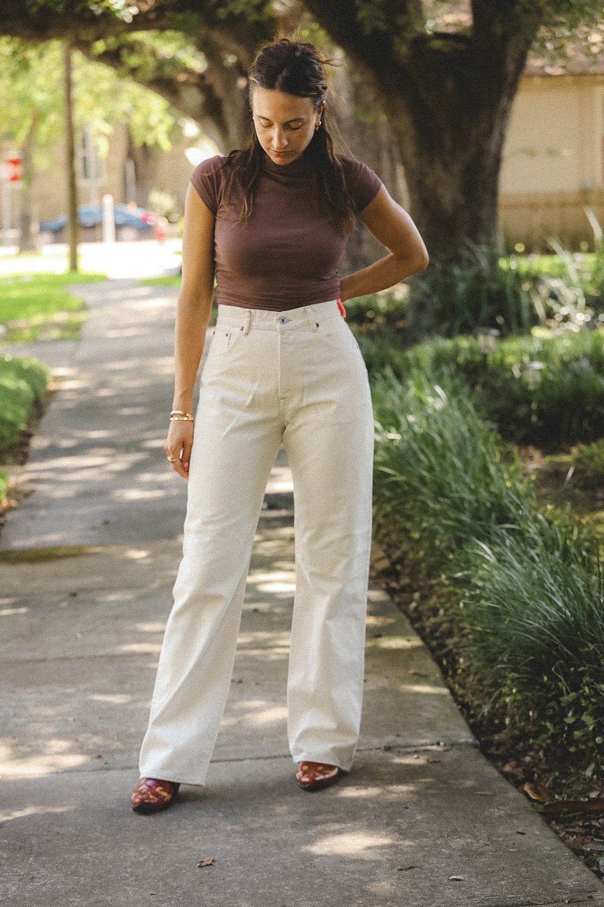 A woman stands on a sunlit sidewalk outdoors, wearing a fitted short-sleeve mauve-brown top and high-waisted off-white wide-leg pants with brown patterned shoes.