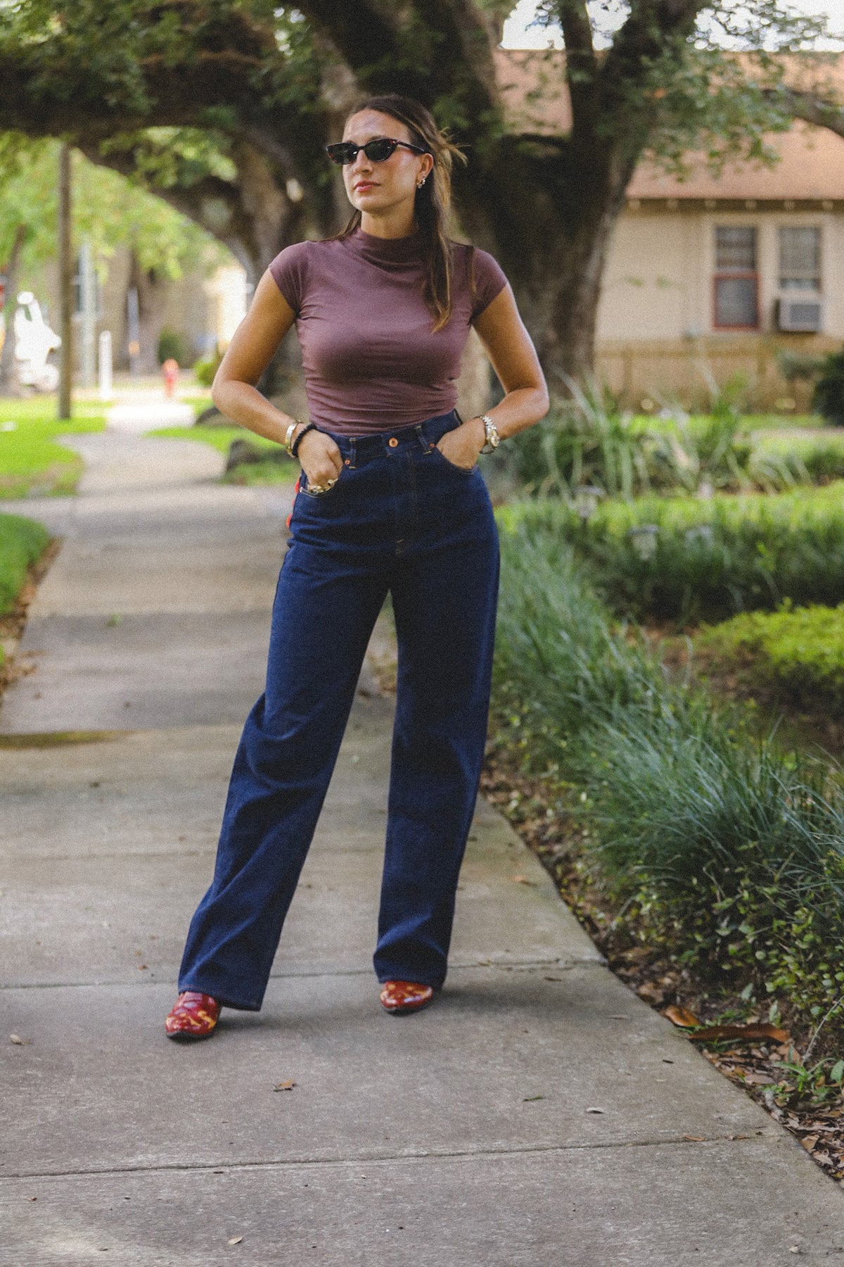 A woman stands on a sidewalk in an outdoor park setting, wearing a fitted mauve short-sleeve top, dark blue wide-leg jeans, and red shoes.
