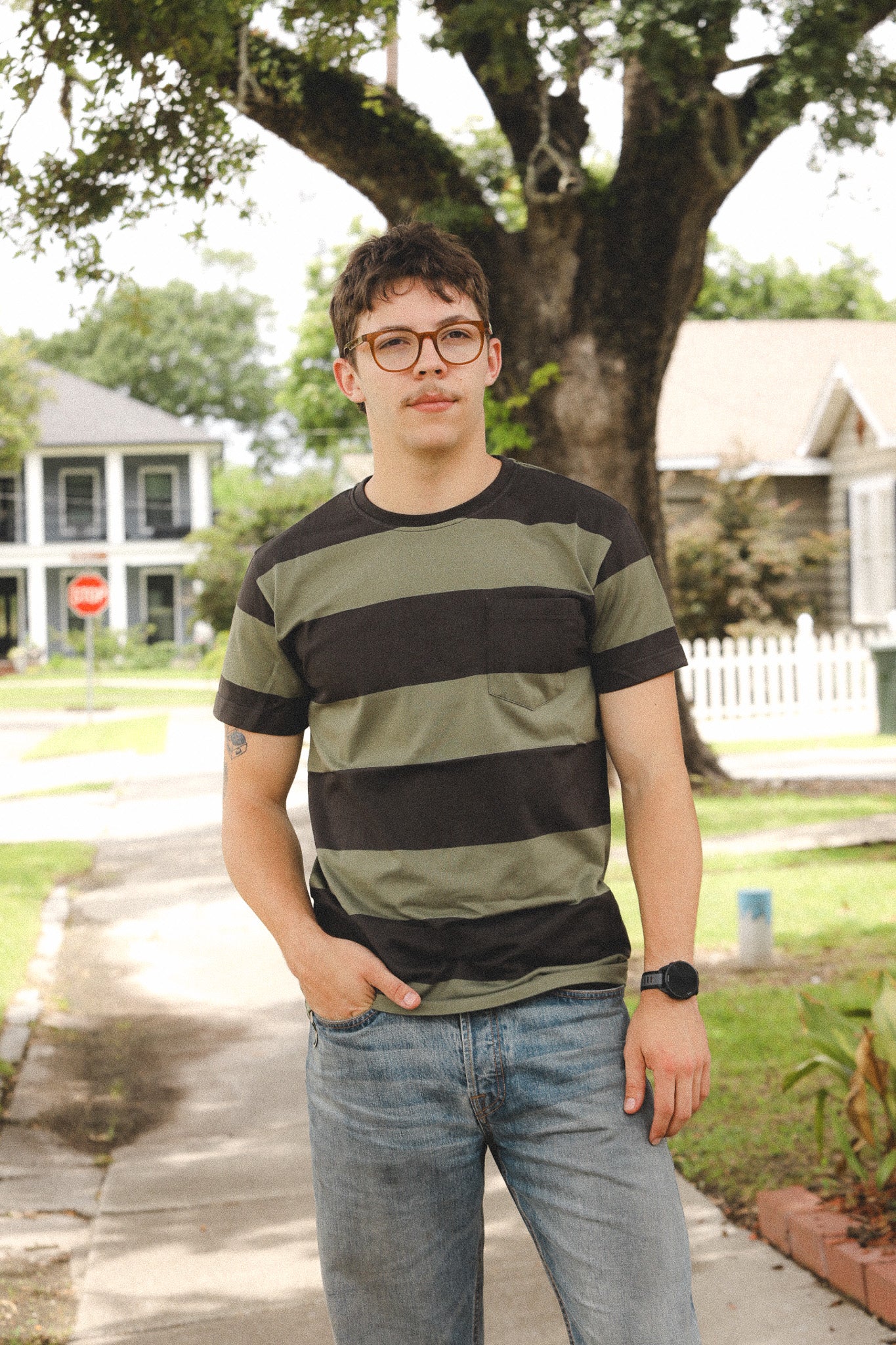 Person wearing a striped shirt standing outdoors with trees and houses in the background