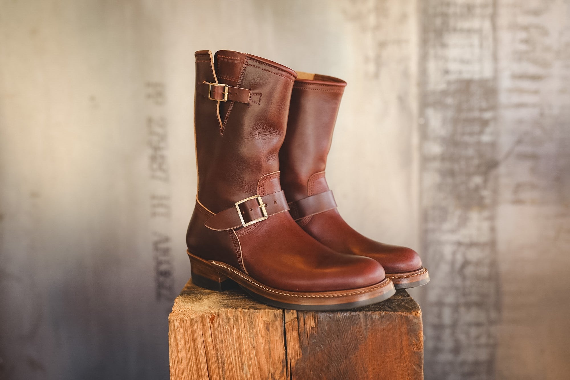 A pair of mid-calf brown leather boots with buckle straps, displayed on a rustic wooden block against a textured, warm-toned background.