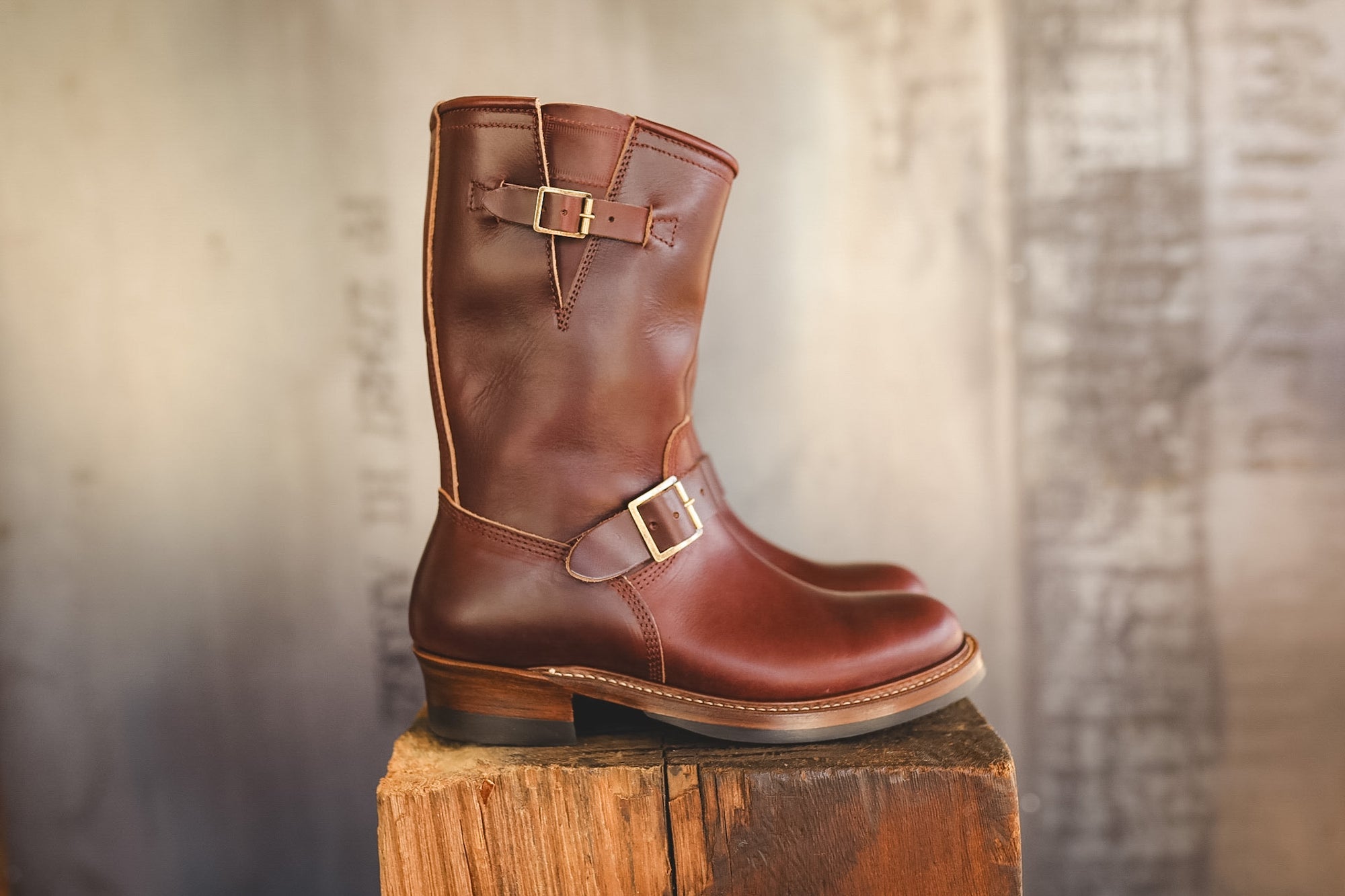 A mid-calf brown leather boot with double buckle straps, displayed on a rustic wooden block against a blurred gray background.