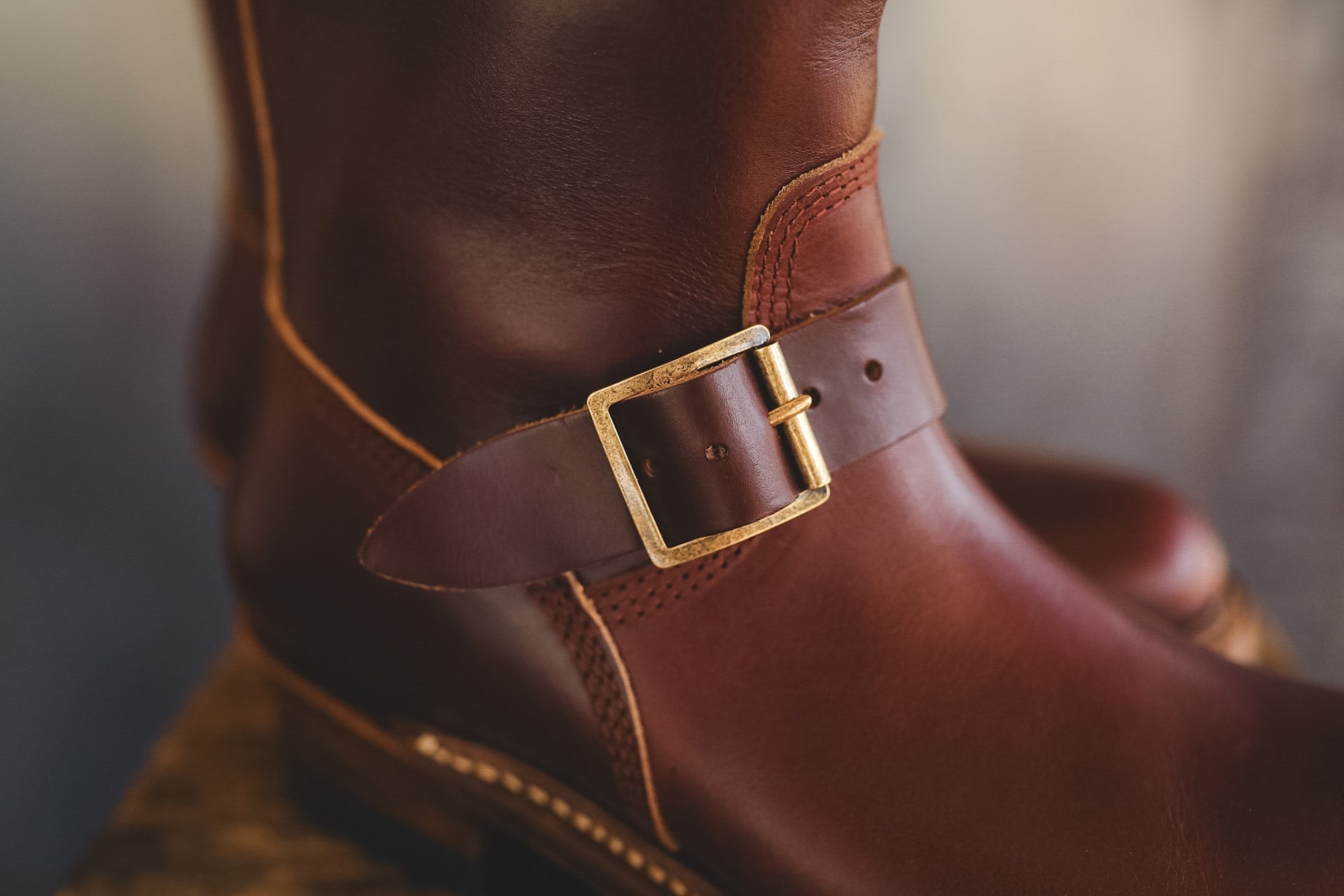 A close-up of a dark brown leather boot featuring a decorative ankle strap with a gold-tone buckle.