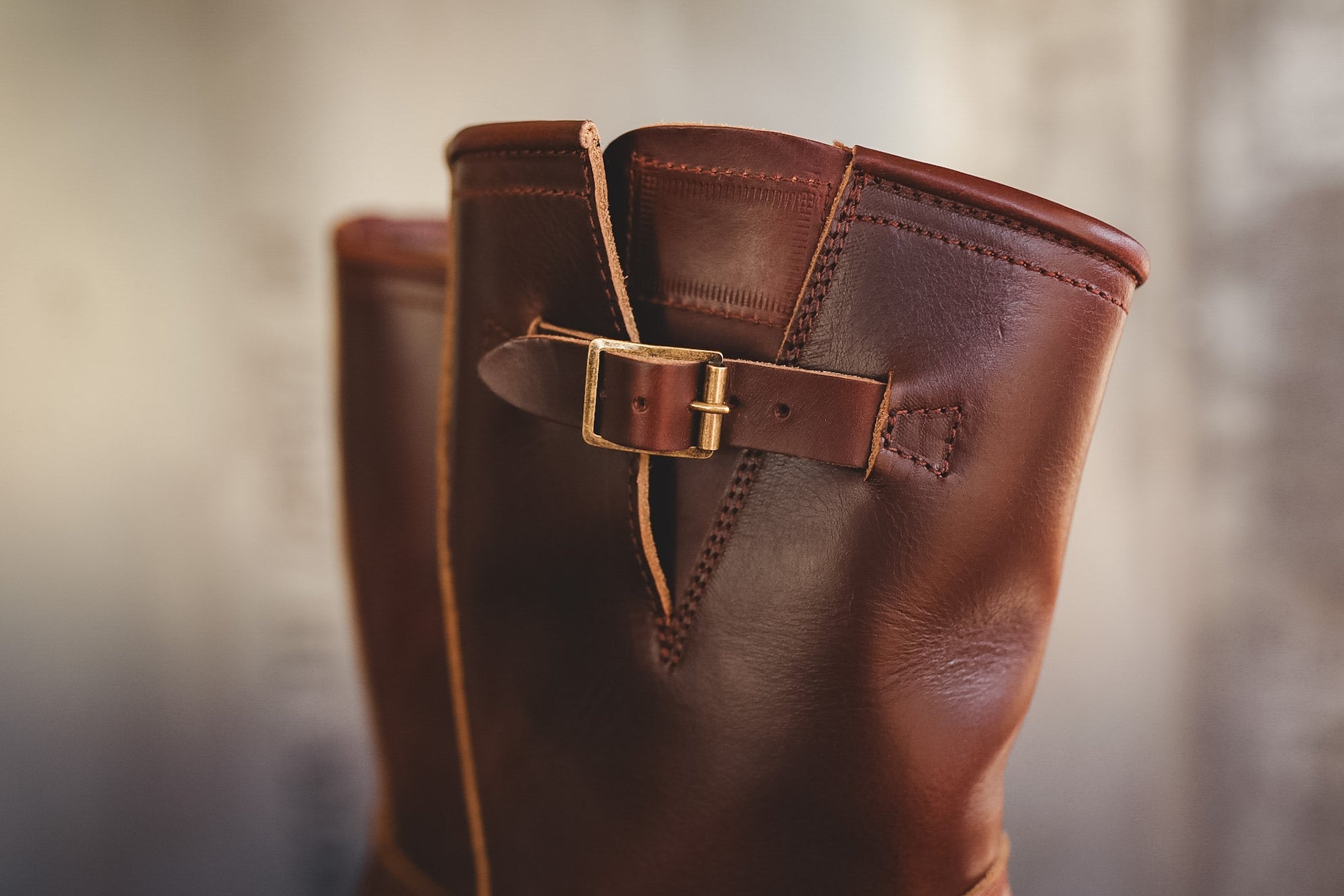 A close-up of a brown leather boot with a decorative strap and brass buckle, photographed against a softly blurred neutral background.