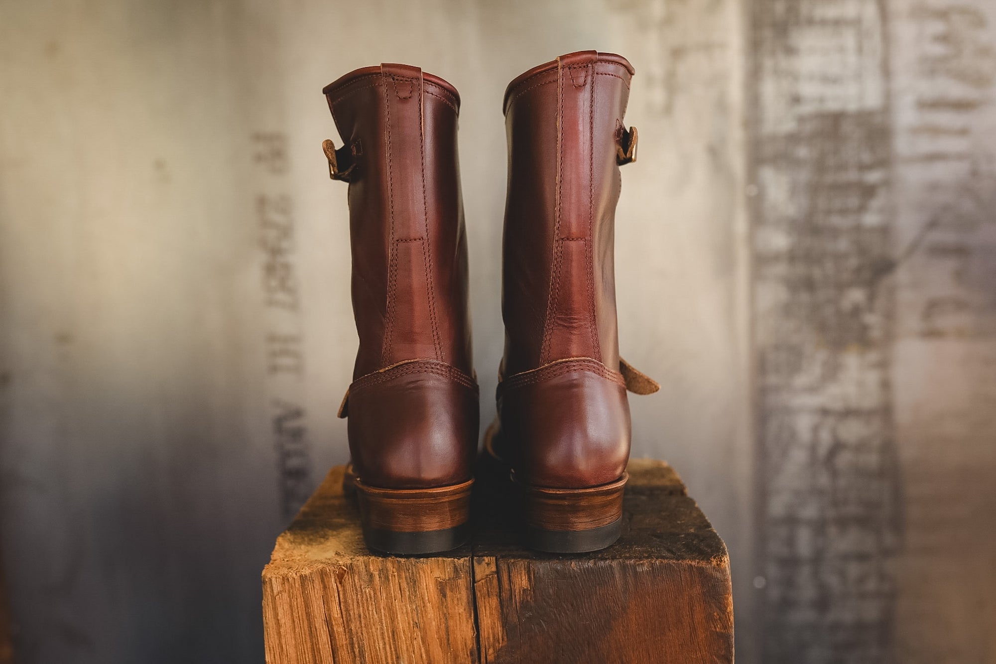 A pair of dark brown leather mid-calf boots with side buckle details, standing side-by-side on a rustic wooden block against a textured, warm-toned background.