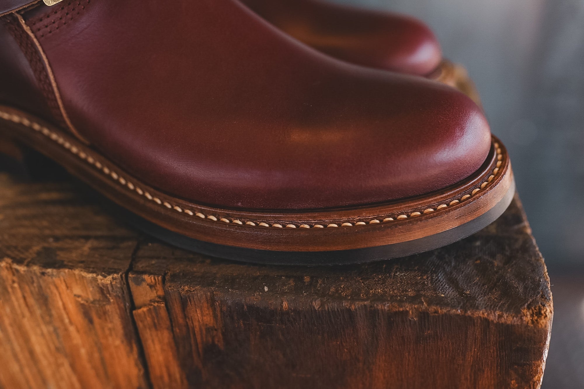 A close-up of a reddish-brown leather boot resting on a rustic wooden stump, showing the smooth leather upper, decorative stitching, and part of the sole.