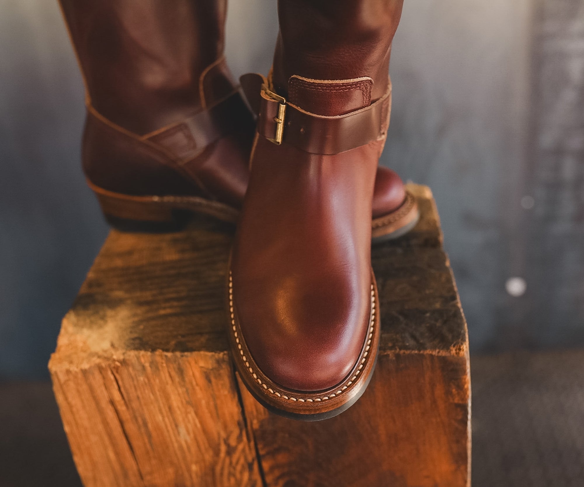 A pair of dark brown leather boots with a buckle strap, shown on a rustic wooden block against a dark gray background.
