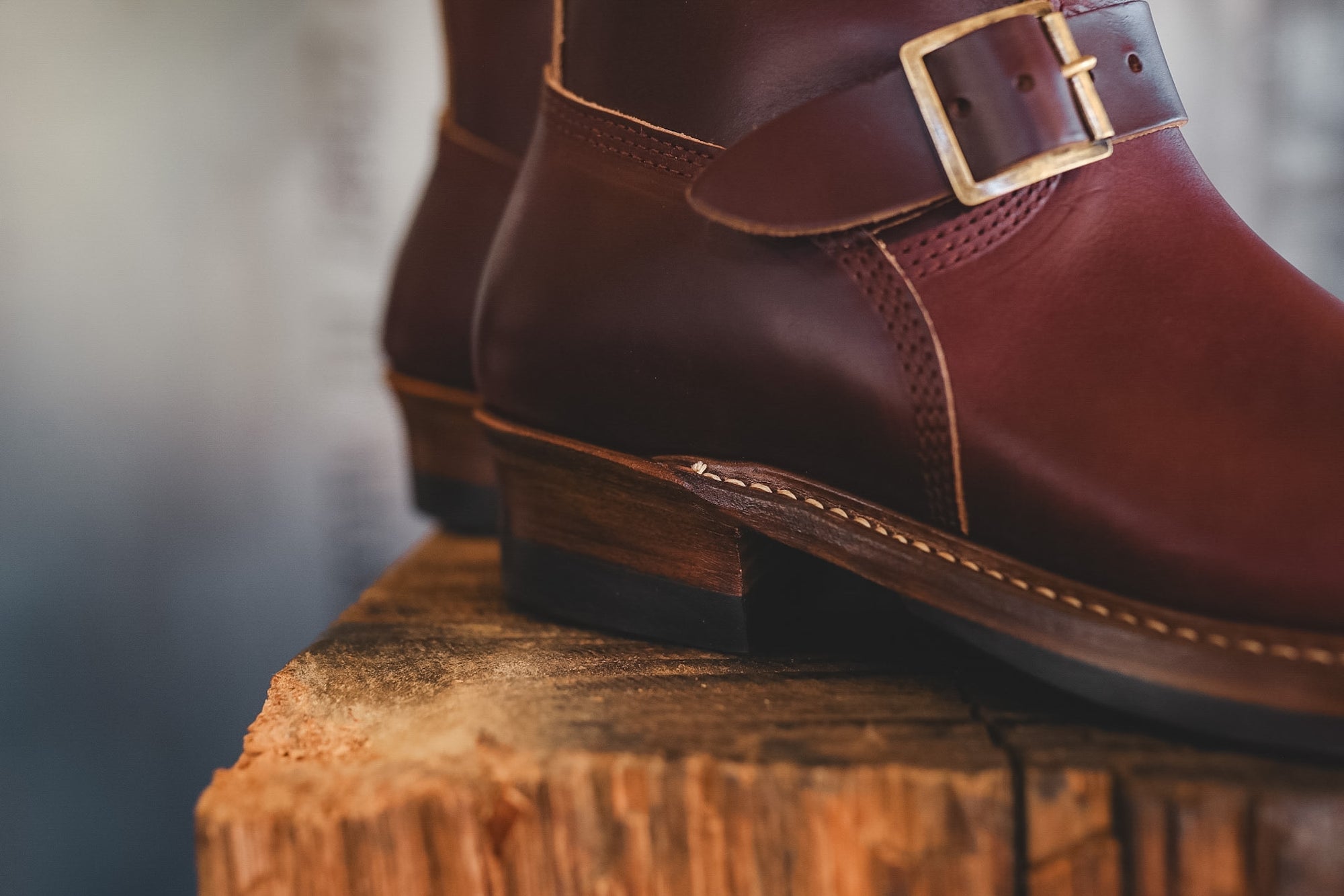 A close-up of dark reddish-brown leather ankle boots with a buckle strap, resting on a rustic wooden log.