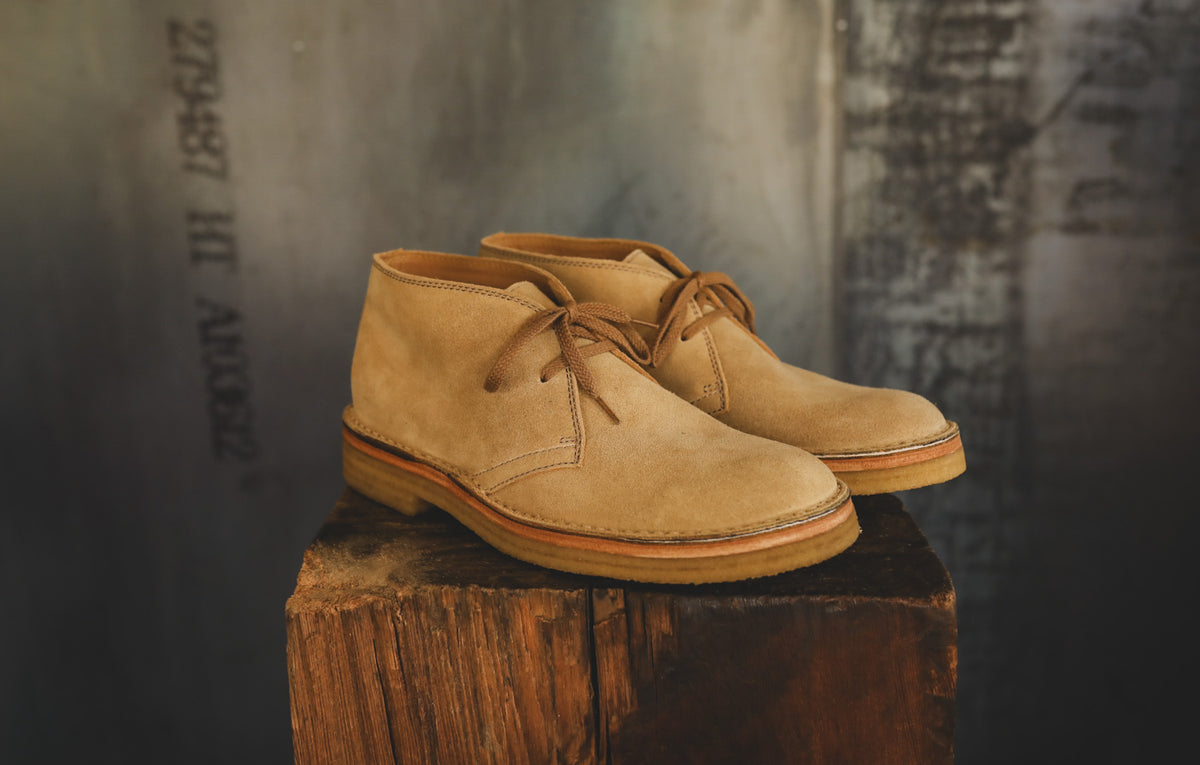 A pair of tan suede Chelsea-style boots with brown laces and a gum sole, displayed on a rustic wooden block against a dark textured background.