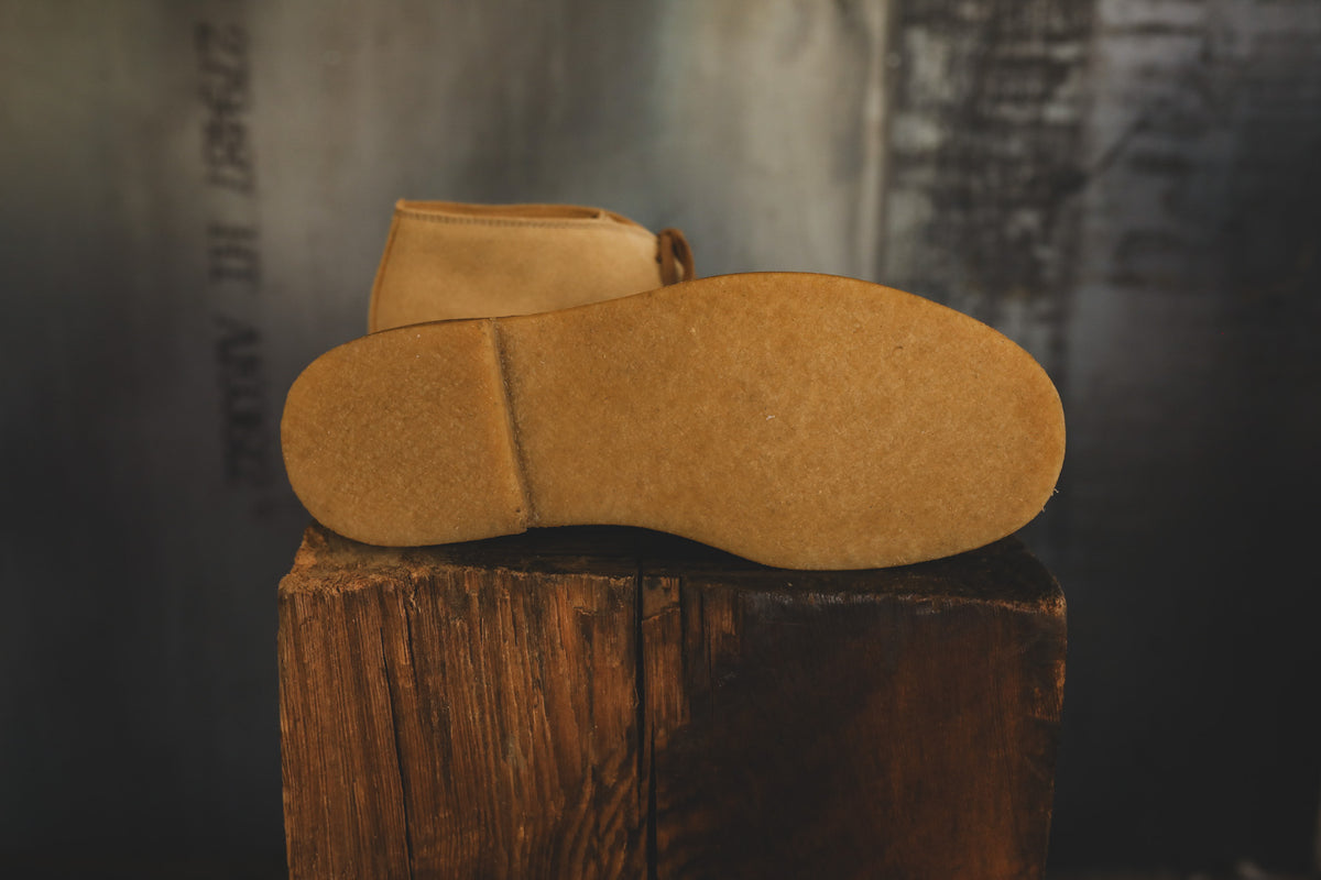 A pair of tan suede boots shown from the sole side, resting on a rustic wooden block against a dark, textured background.