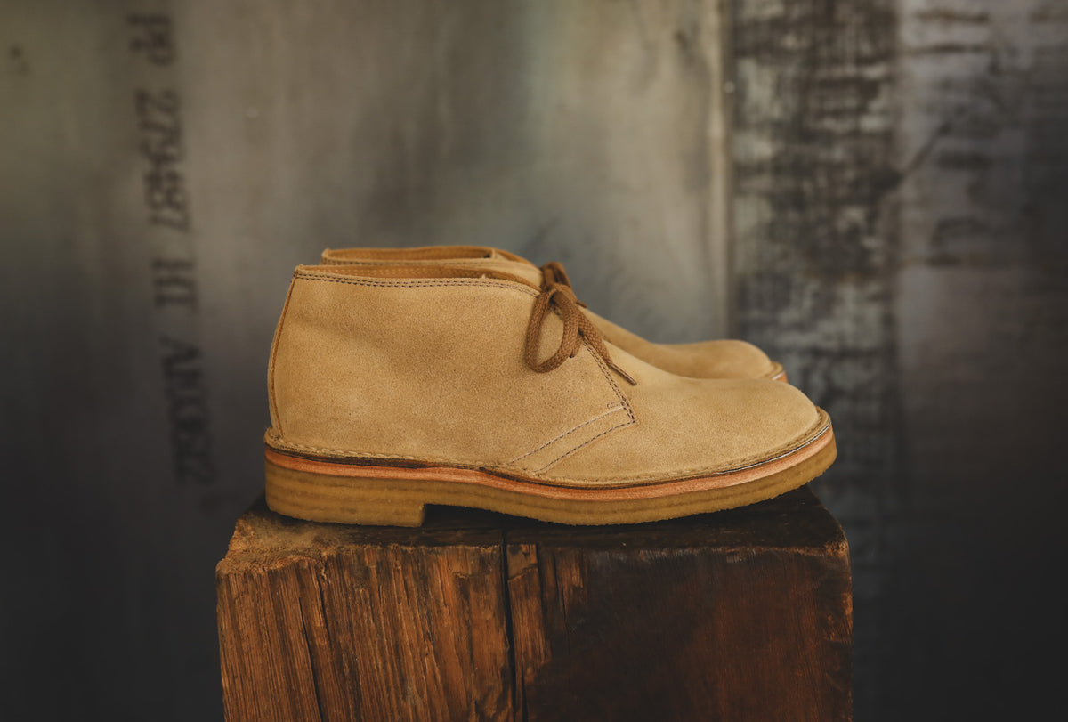 A pair of tan suede chukka boots with brown laces, displayed on a rustic wooden block against a dark, textured industrial background.