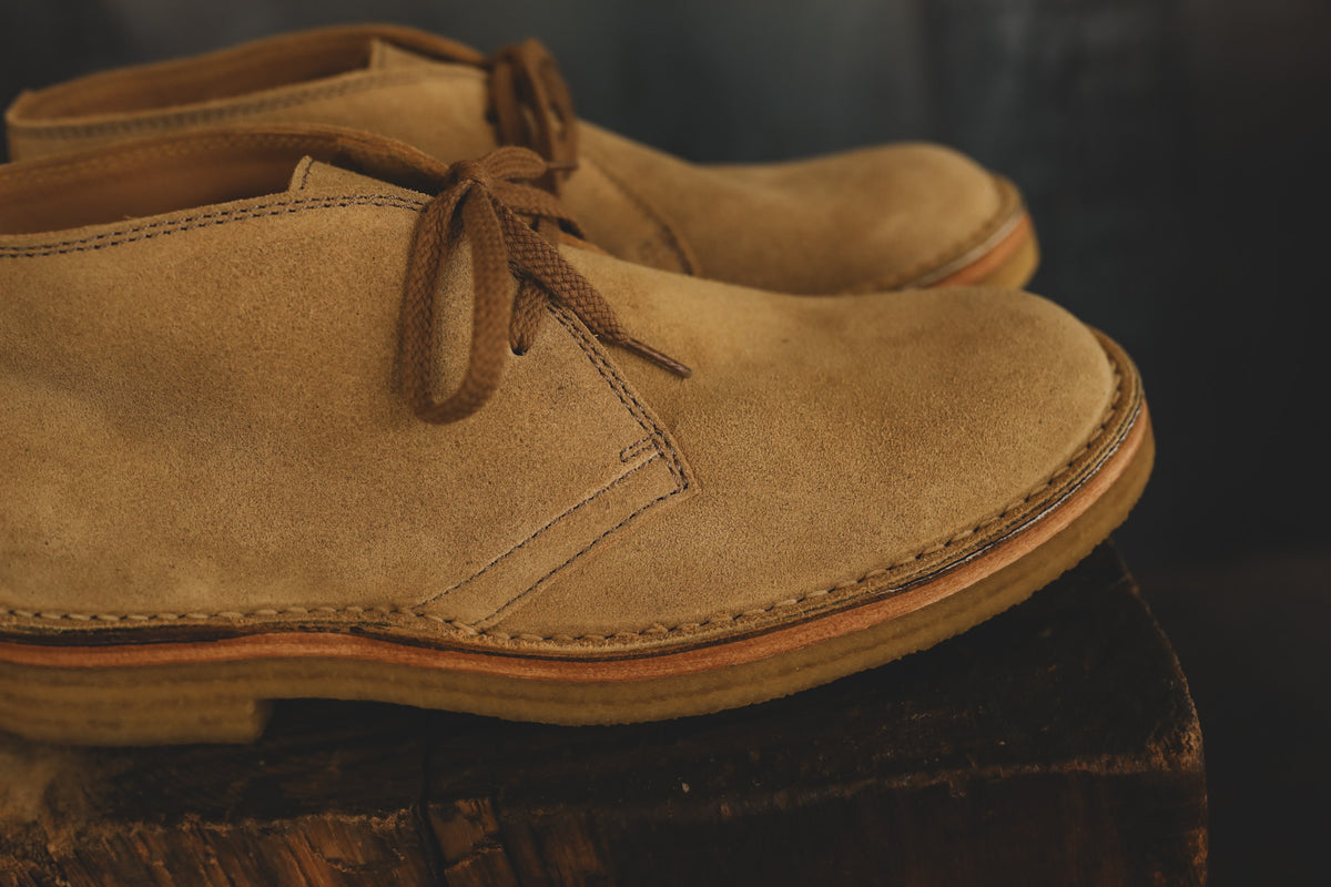 A pair of tan suede lace-up ankle boots resting on a rustic wooden block against a dark, softly blurred background.