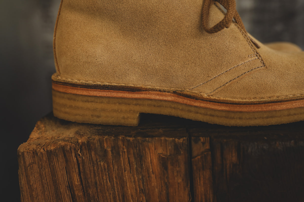 A close-up side view of a tan suede lace-up boot resting on a rustic wooden stump against a dark blurred background.
