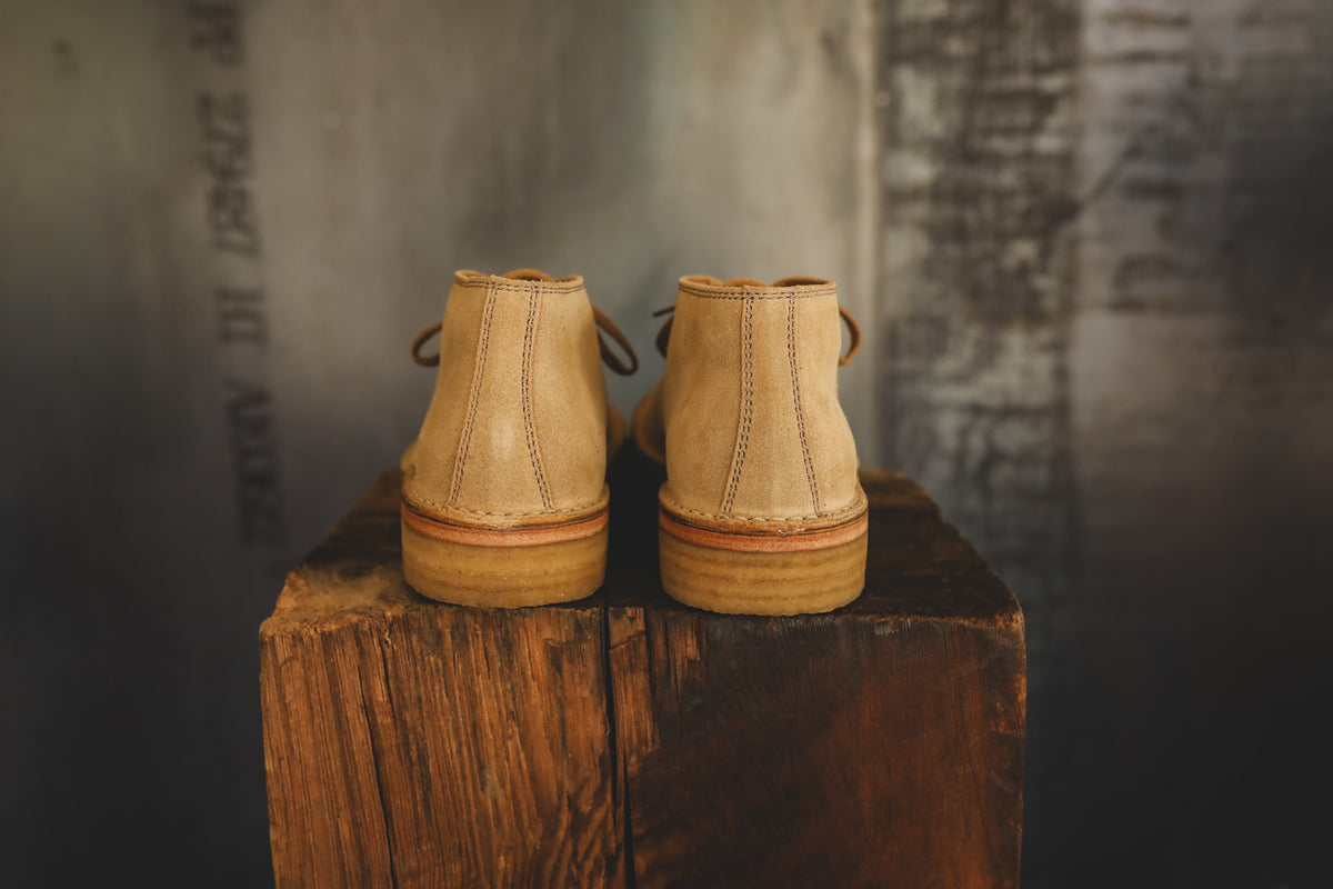 A pair of light tan leather lace-up ankle boots shown from the back, resting on a rustic wooden block against a dark textured studio background.