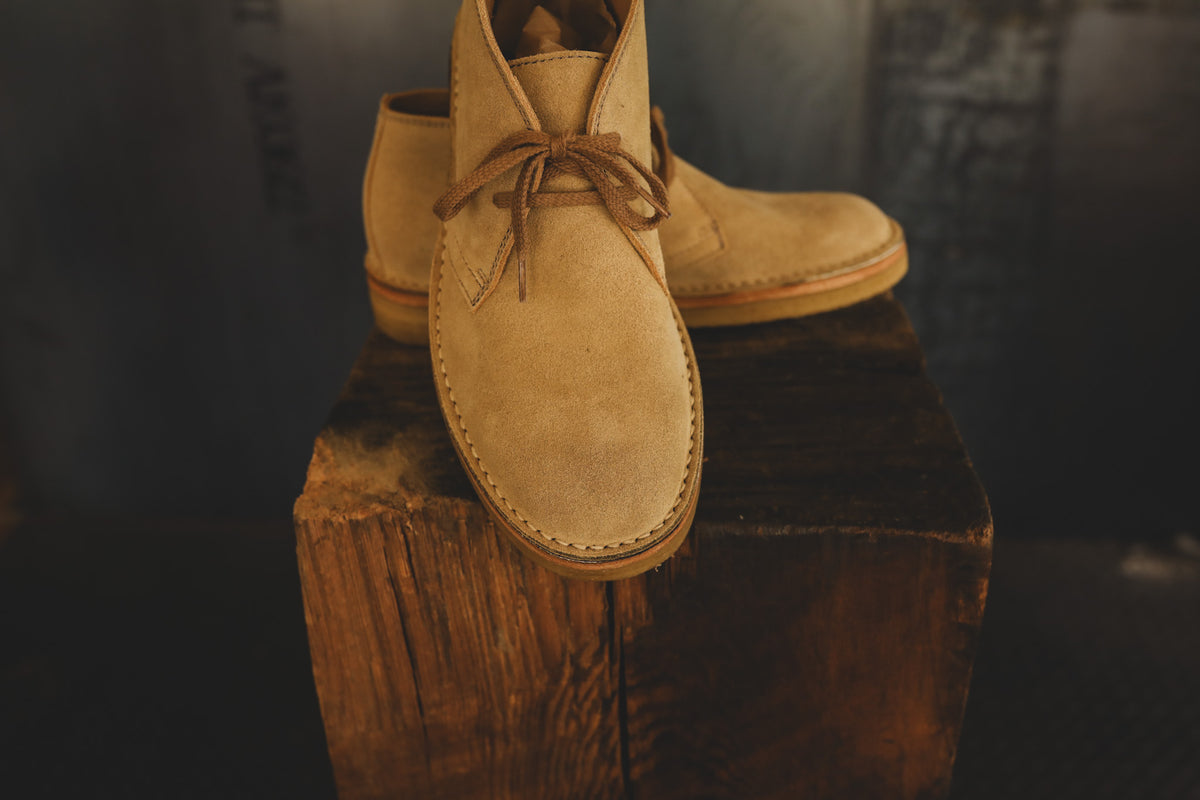 A pair of tan suede lace-up ankle boots displayed on a rustic wooden block against a dark, moody background.