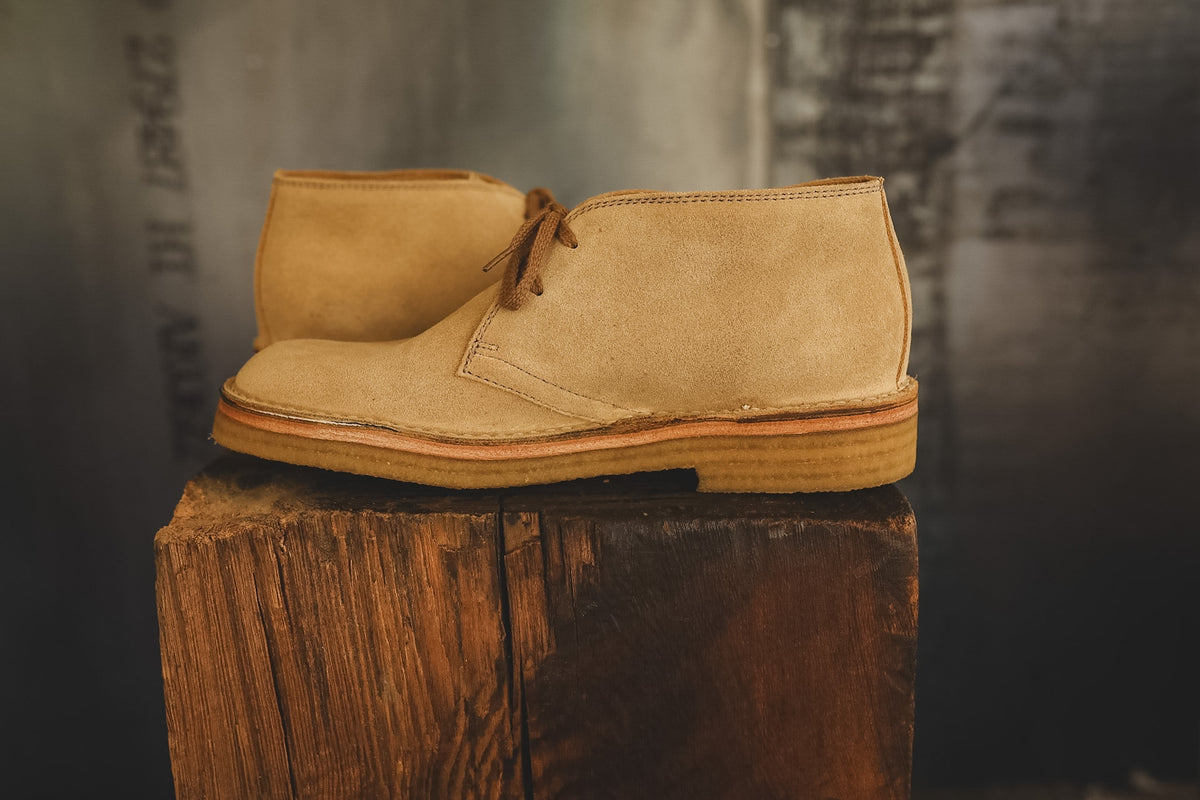 A pair of tan suede chukka boots with brown laces, displayed on a rustic wooden block against a dark, textured background.