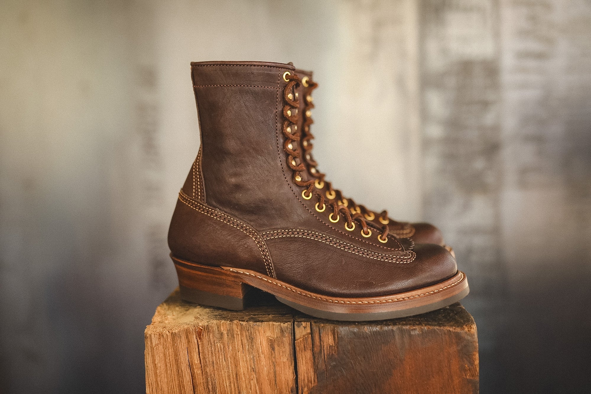A pair of dark brown leather lace-up ankle boots with gold-tone eyelets and stacked brown heels, displayed on a rustic wooden block against a blurred gray background.