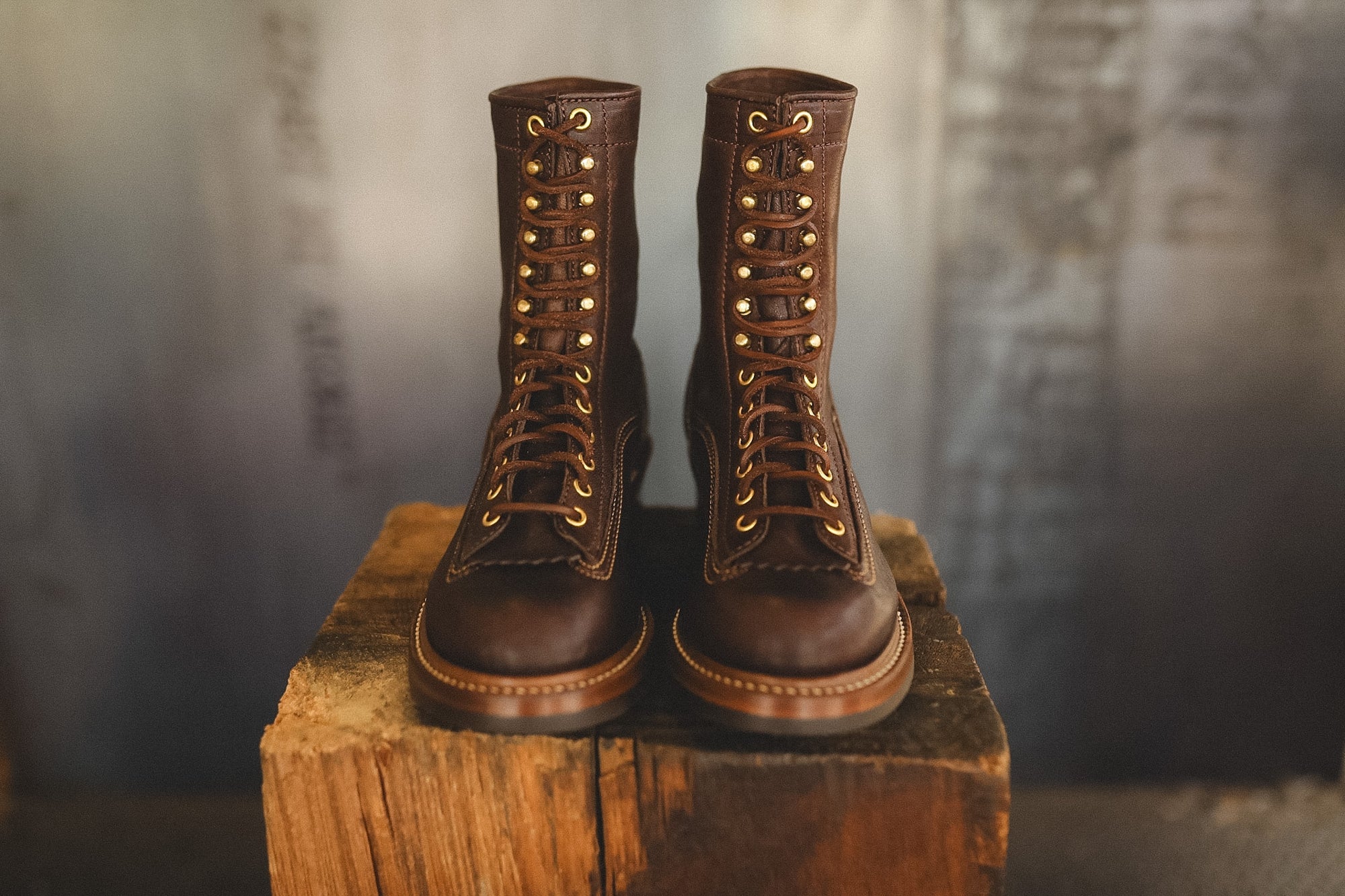 A pair of dark brown leather lace-up boots with brass eyelets and laces, displayed side-by-side on a rustic wooden block against a blurred gray background.