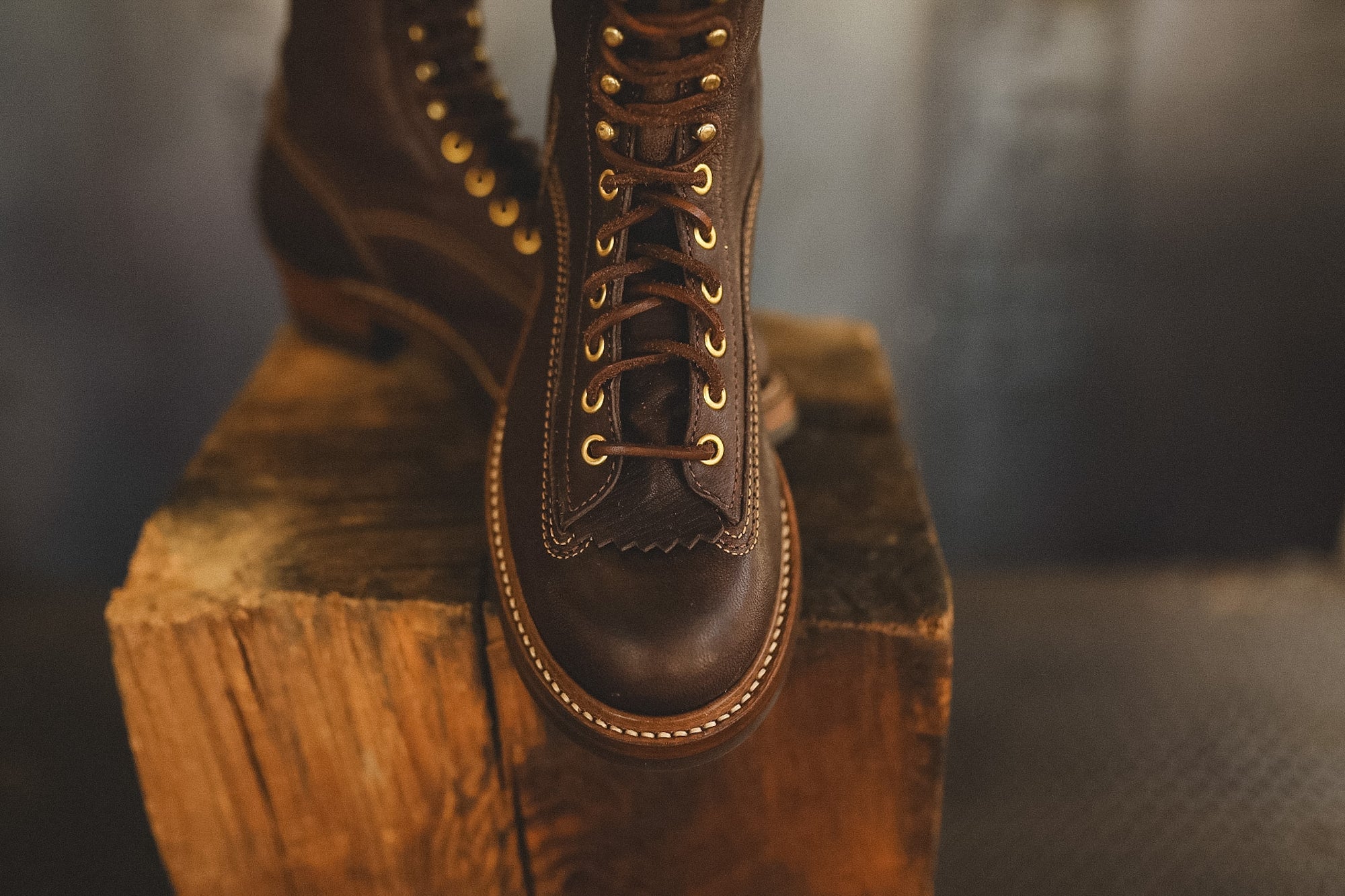 A pair of dark brown leather lace-up boots with gold-tone eyelets, displayed on a rustic wooden block against a dark, softly blurred background.