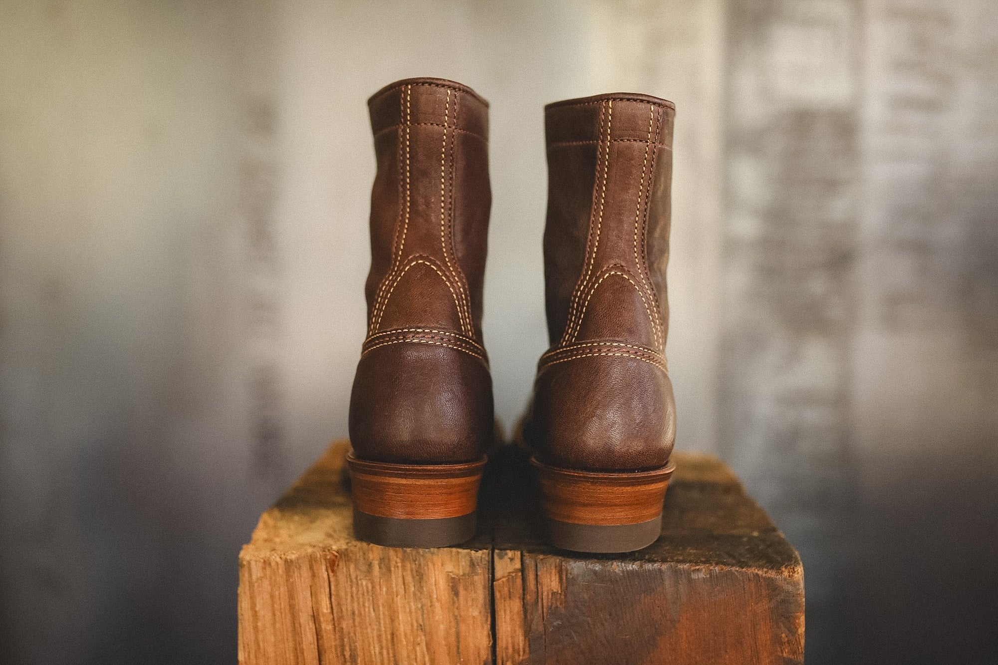 A pair of dark brown leather ankle boots with light tan contrast stitching, displayed from the back on a rustic wooden block against a blurred gray background.