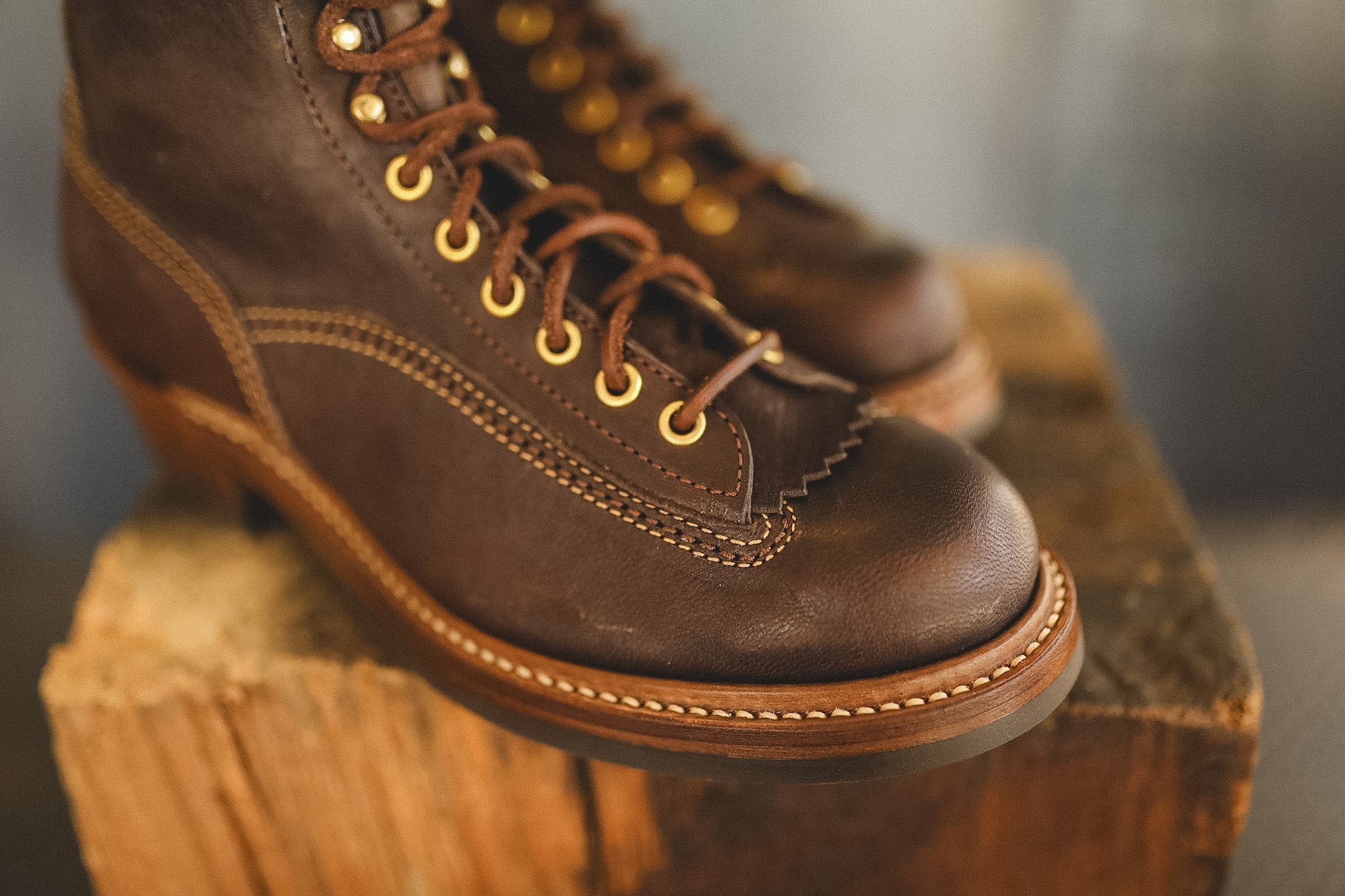 A close-up of a dark brown leather lace-up boot with gold eyelets and contrast stitching, resting on a rustic wooden block.