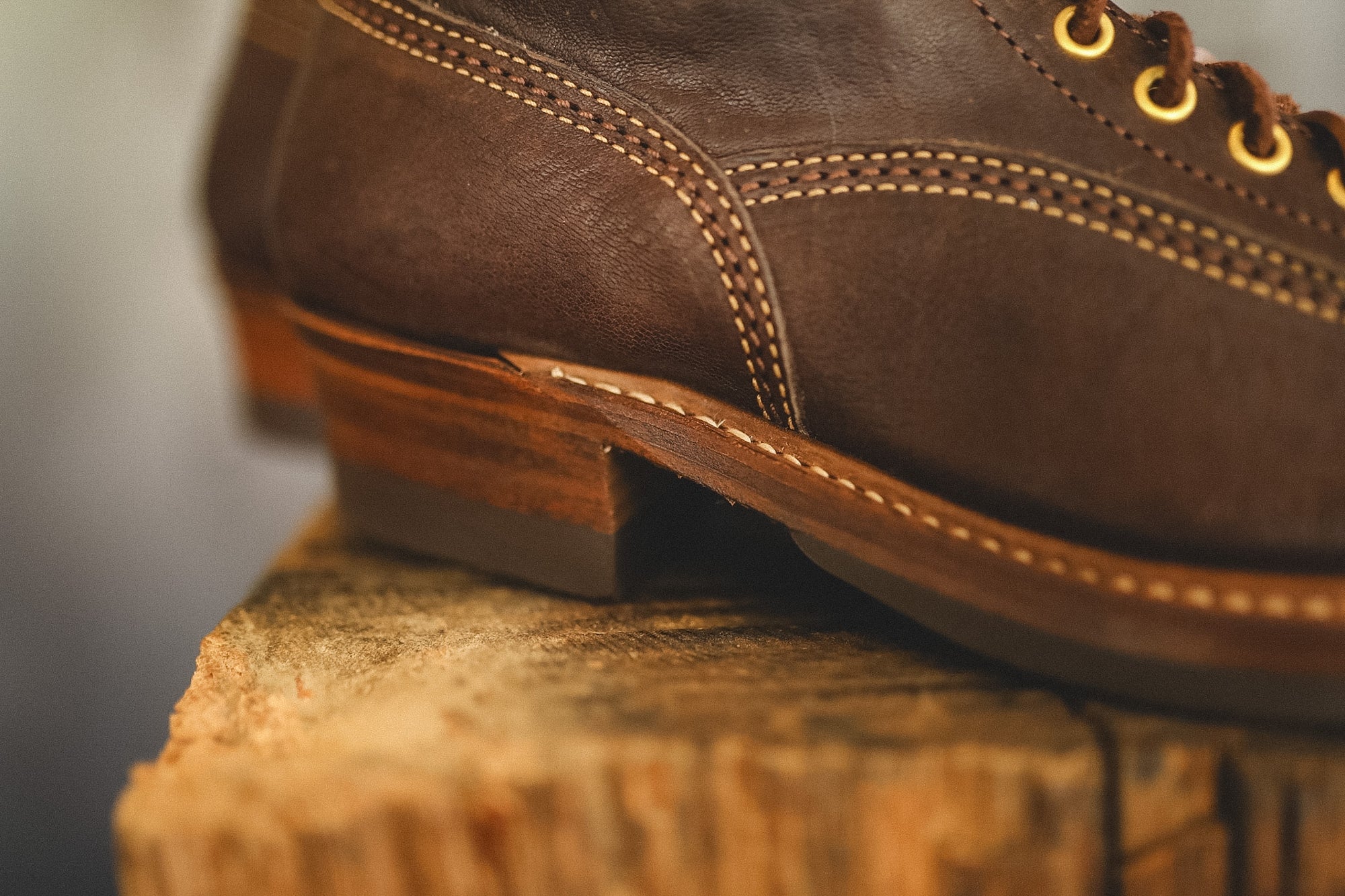 A close-up shot of a dark brown leather lace-up boot resting on a rustic wooden log, showing the stitching, heel, and eyelets in sharp detail against a blurred background.