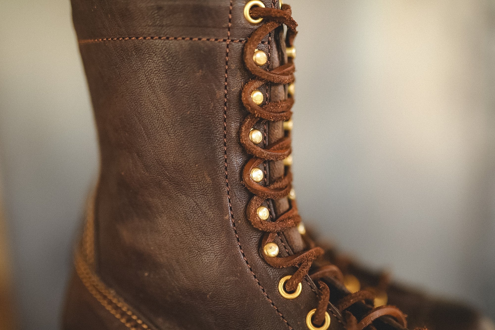 Close-up of a dark brown leather lace-up boot with brass eyelets and laces, shown against a softly blurred neutral background.