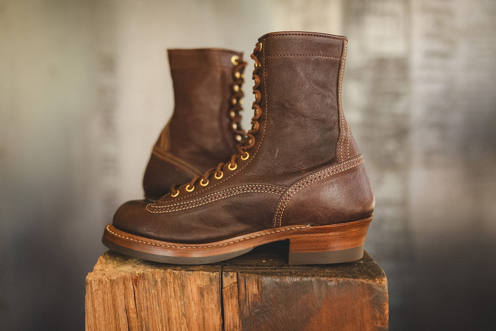 A pair of dark brown leather lace-up boots with gold-toned metal eyelets and stacked wooden heels, displayed on a rustic wooden block against a blurred gray background.