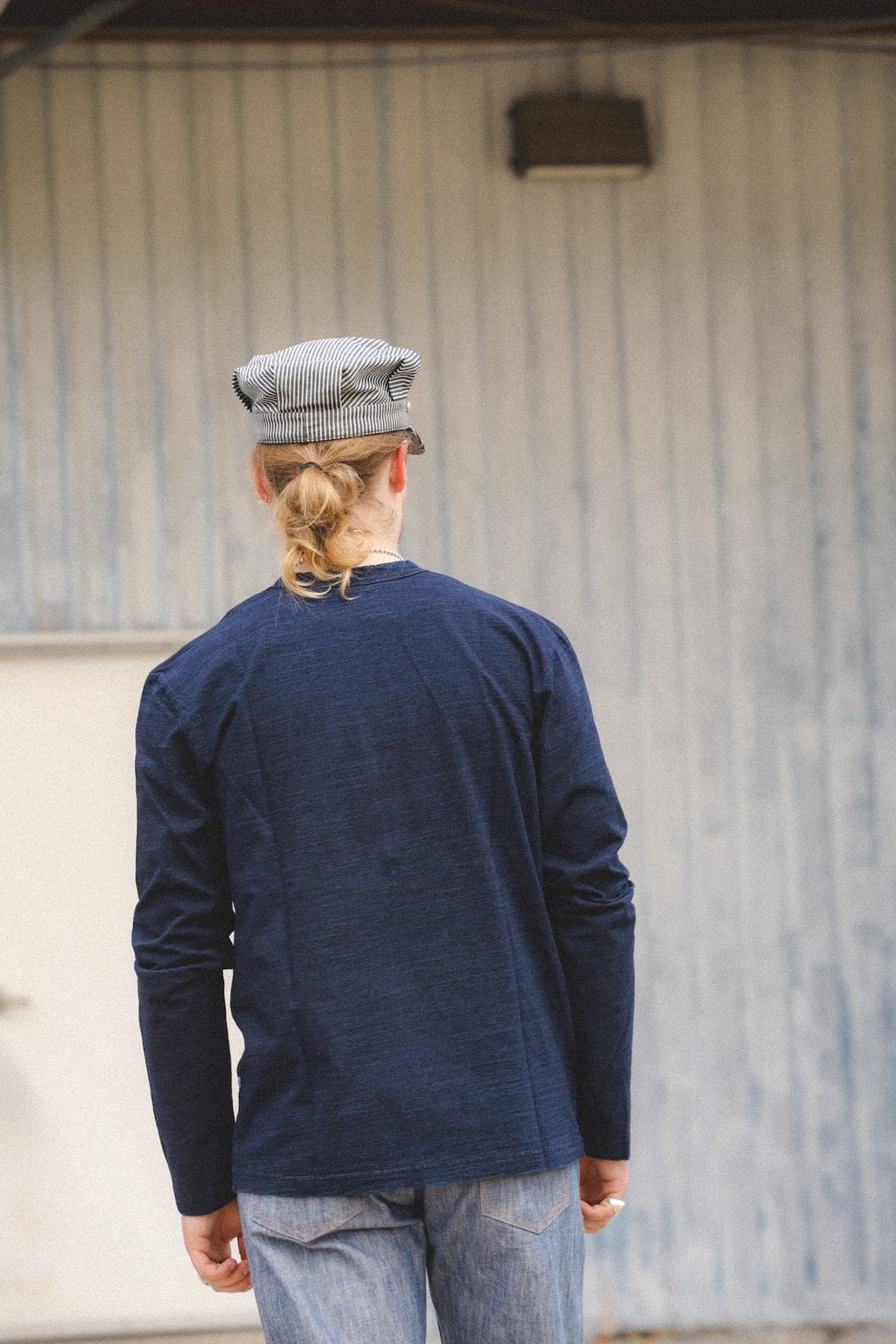A man viewed from behind wearing a navy blue long-sleeve shirt, a gray patterned flat cap, and light blue jeans, standing in front of a gray corrugated metal wall.