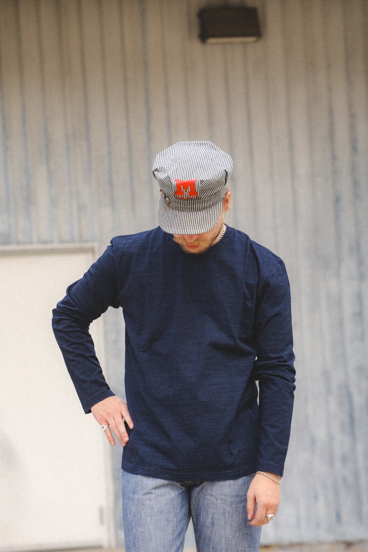 A man wearing a navy blue long-sleeve shirt, light blue jeans, and a gray striped flat cap with a red logo, standing in front of a gray corrugated metal wall.