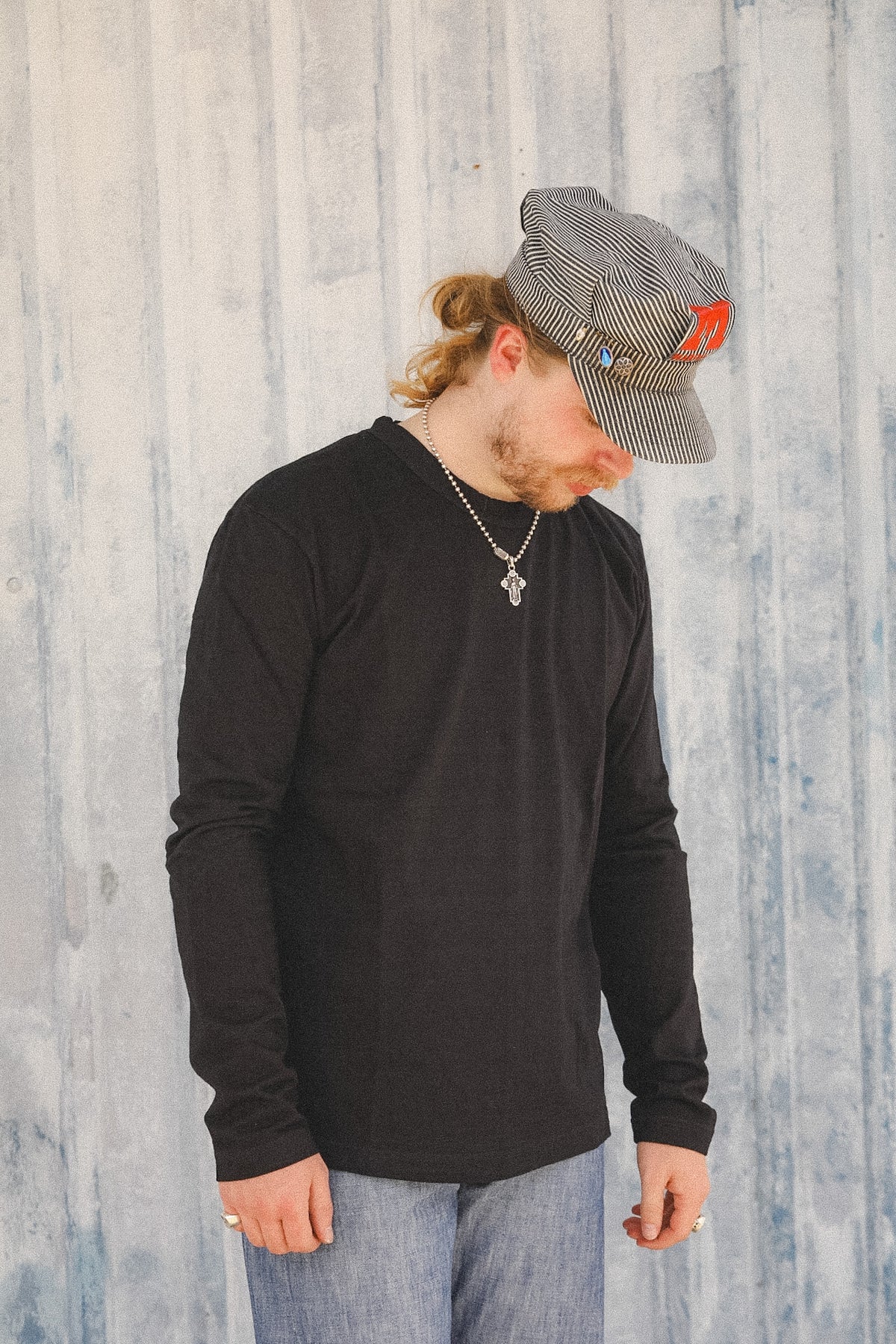 A man wearing a black long-sleeve crewneck shirt, blue jeans, a silver chain necklace, and a gray and white striped cap with a red logo, standing against a light blue-gray wood-paneled wall.