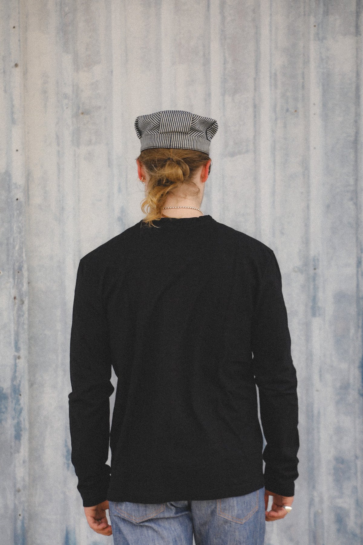A person viewed from behind wearing a black long-sleeve shirt, blue jeans, and a gray and white striped flat cap, standing against a textured gray wall.